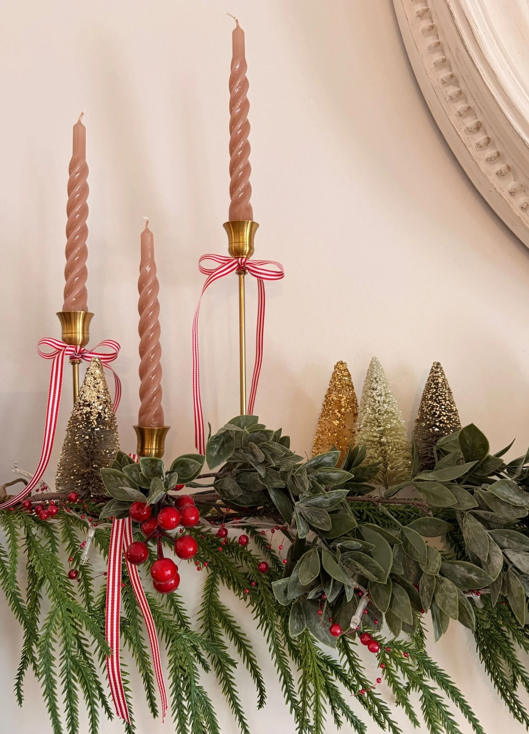 Pink taper candles in brass candle holders with red ribbon bows, evergreen branches, red berries and bottle brush Christmas trees.