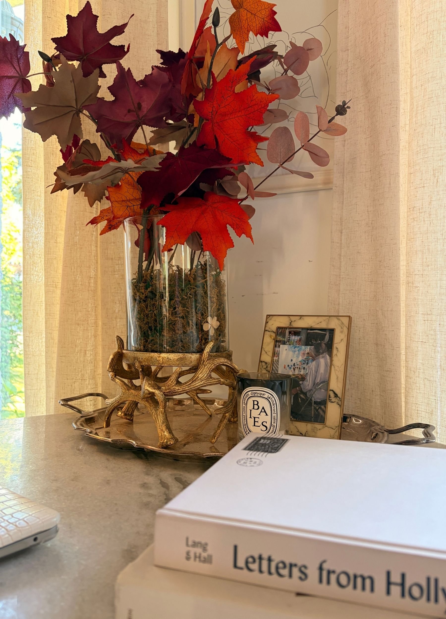 Soft fall decor with colourful autumn leaves arranged in a glass vase with moss, layered with books, a candle and warm vintage-inspired details.