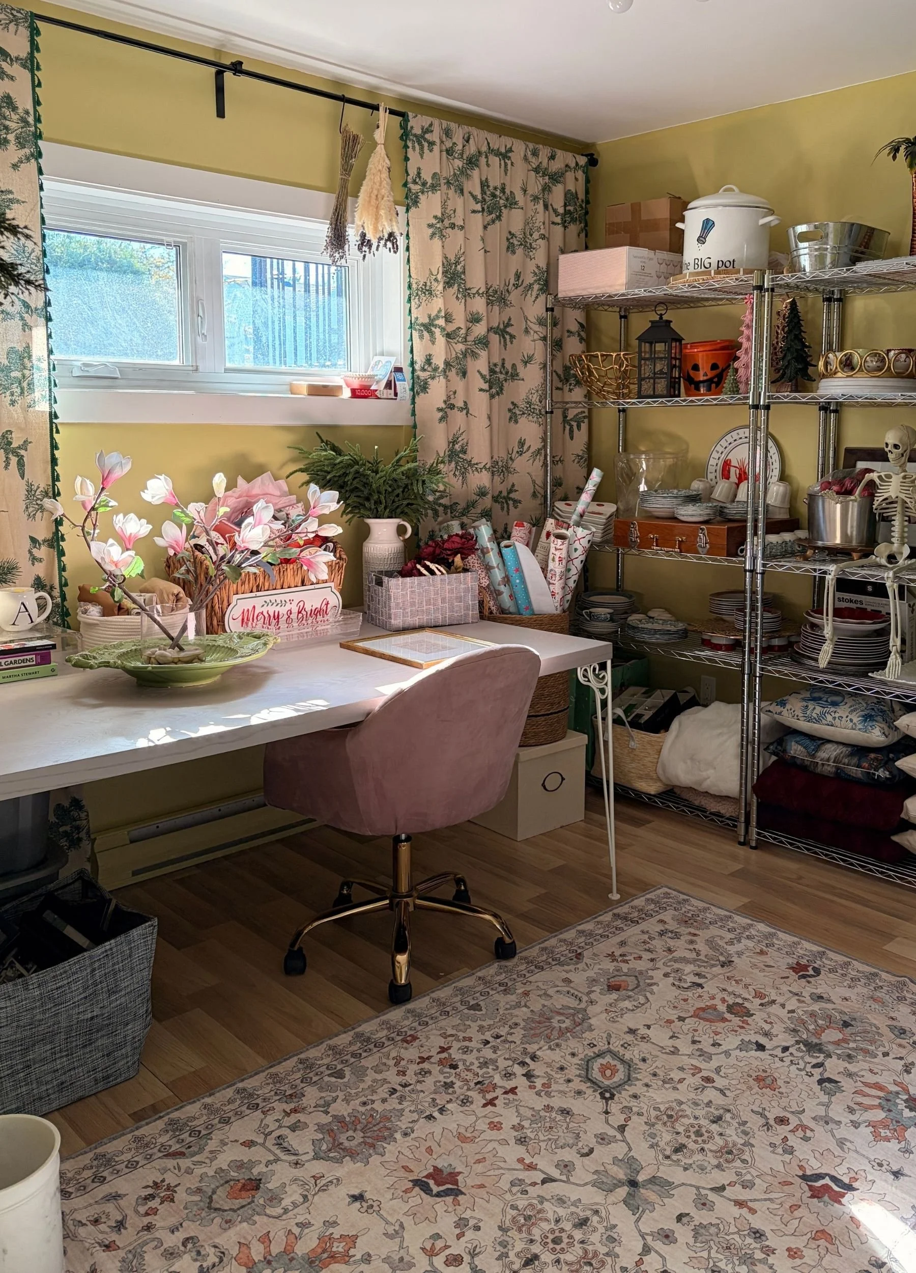 Martha Room craft and holiday storage space with a large worktable, pink chair, patterned rug, open shelving and seasonal decor.