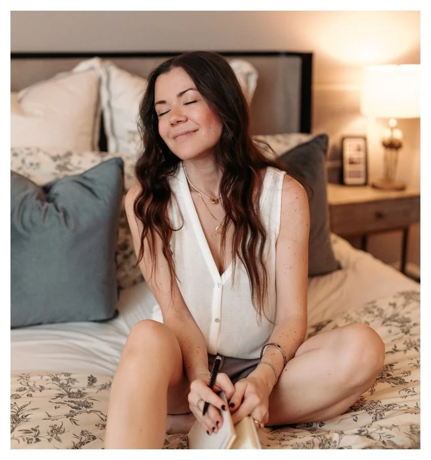 Woman sitting cross-legged on a bed holding a journal in a softly lit bedroom.