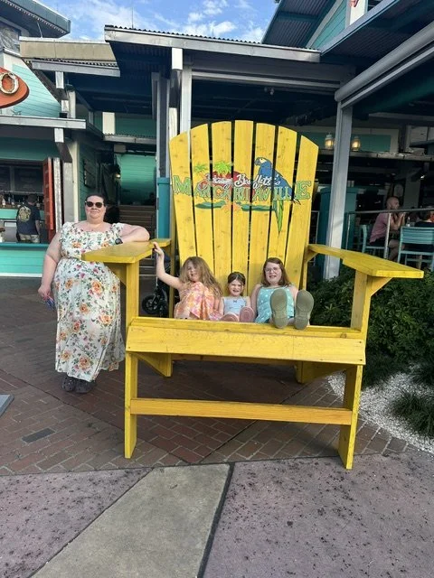 Three children sitting on a large yellow Adirondack chair with the words 'Bai Beach Mocules' painted on the backrest, and an adult woman standing next to it on a brick sidewalk outside a building.
