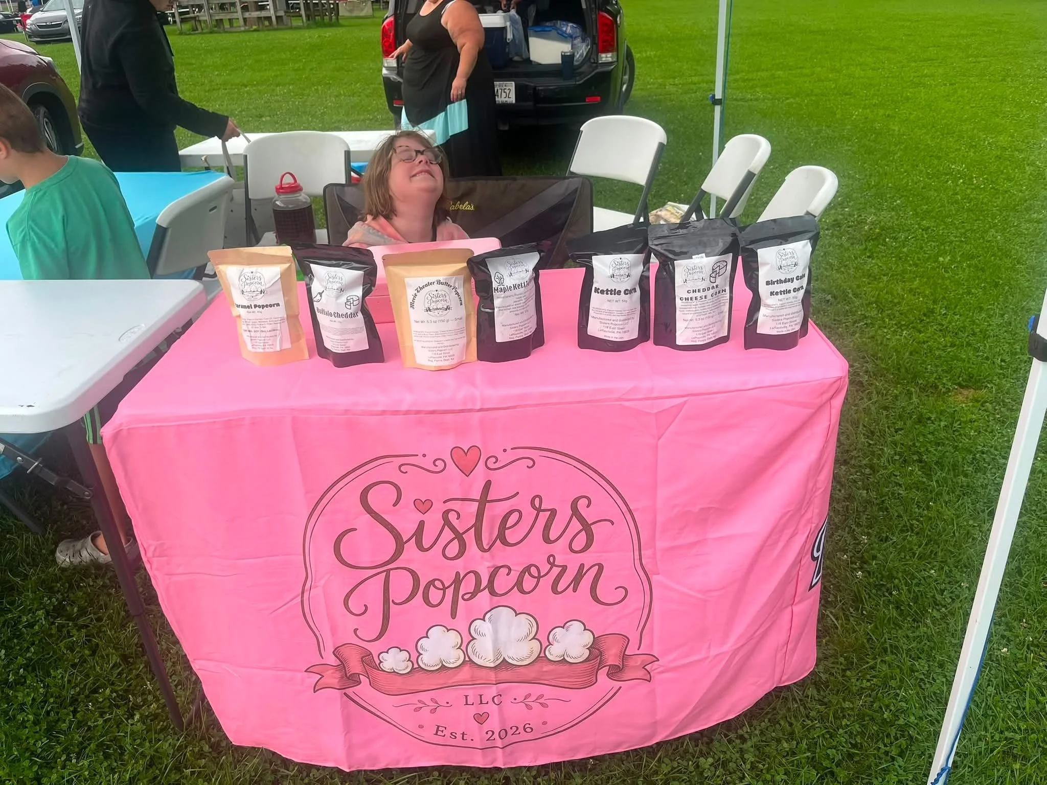 A pink tablecloth with the words 'Sisters in Decor' and illustrations of popcorn on a table at an outdoor event. Several bags of popcorn and a drink bottle are on the table. A person with glasses and a gray hoodie is smiling, sitting behind the table. Other people and cars are visible in the background on green grass.