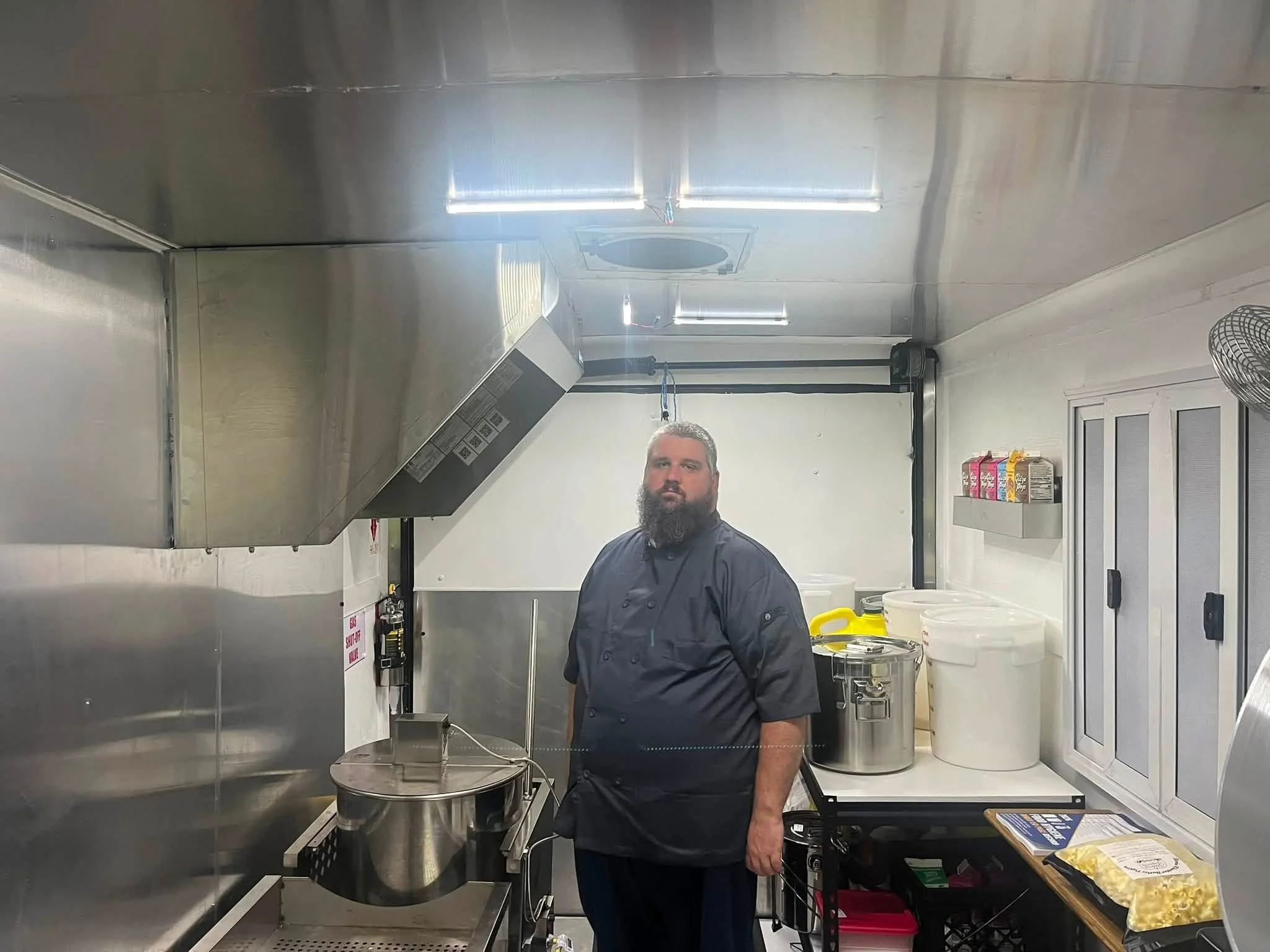 A man with a beard wearing a black chef's uniform standing in a commercial kitchen with stainless steel appliances and containers.