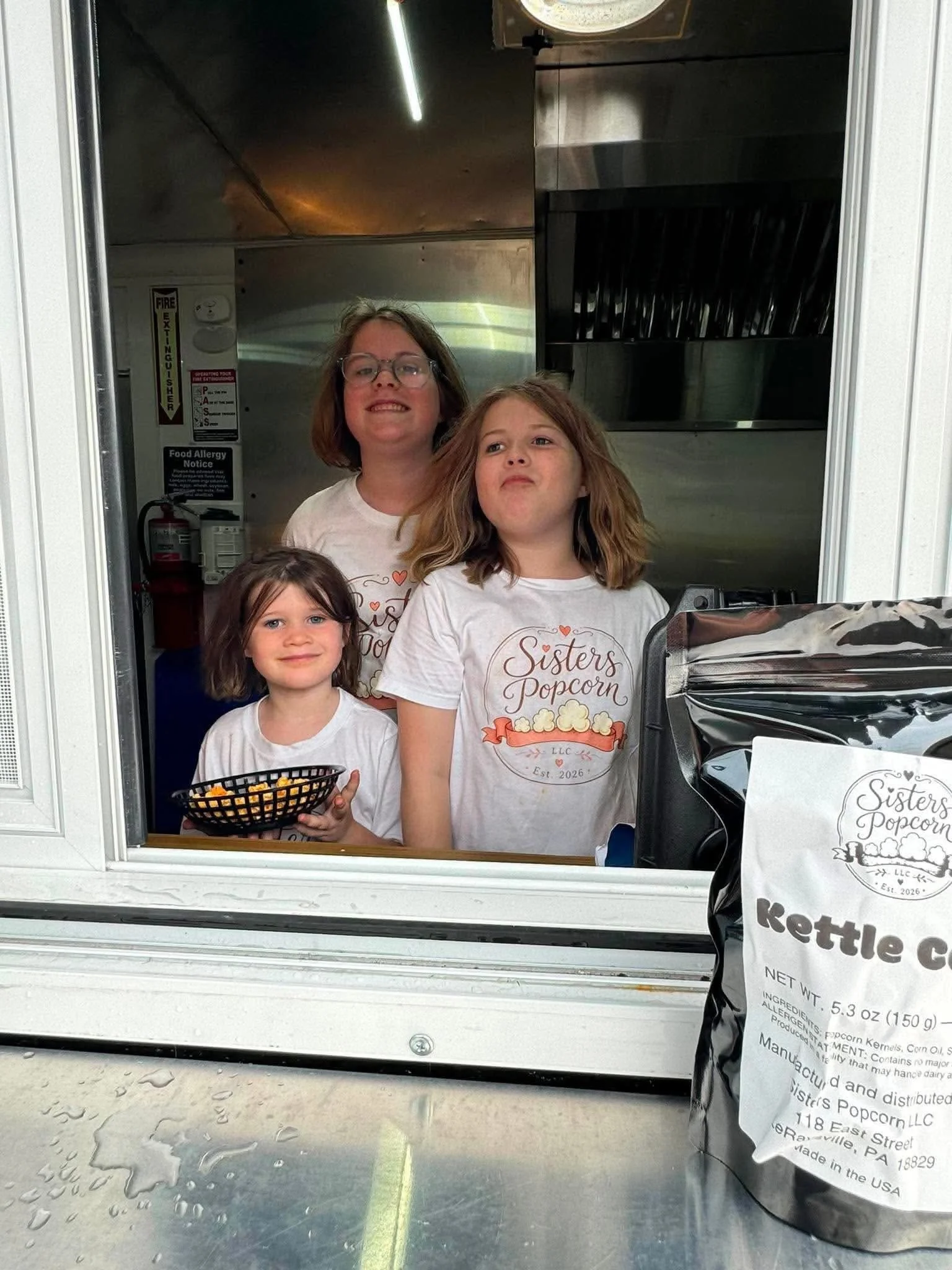 Four young girls inside a popcorn stand window, all wearing matching white shirts with the words "Sisters Popcorn" and a popcorn graphic, smiling and looking outside. One girl in front holds a small basket of popcorn.