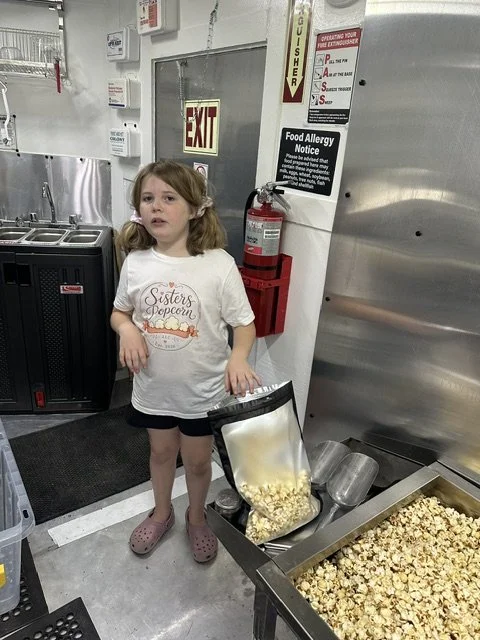 A young girl with pigtails and a white t-shirt with a popcorn design, standing next to a popcorn machine in a building kitchen or snack area. There is a fire extinguisher and safety signs on the wall behind her.