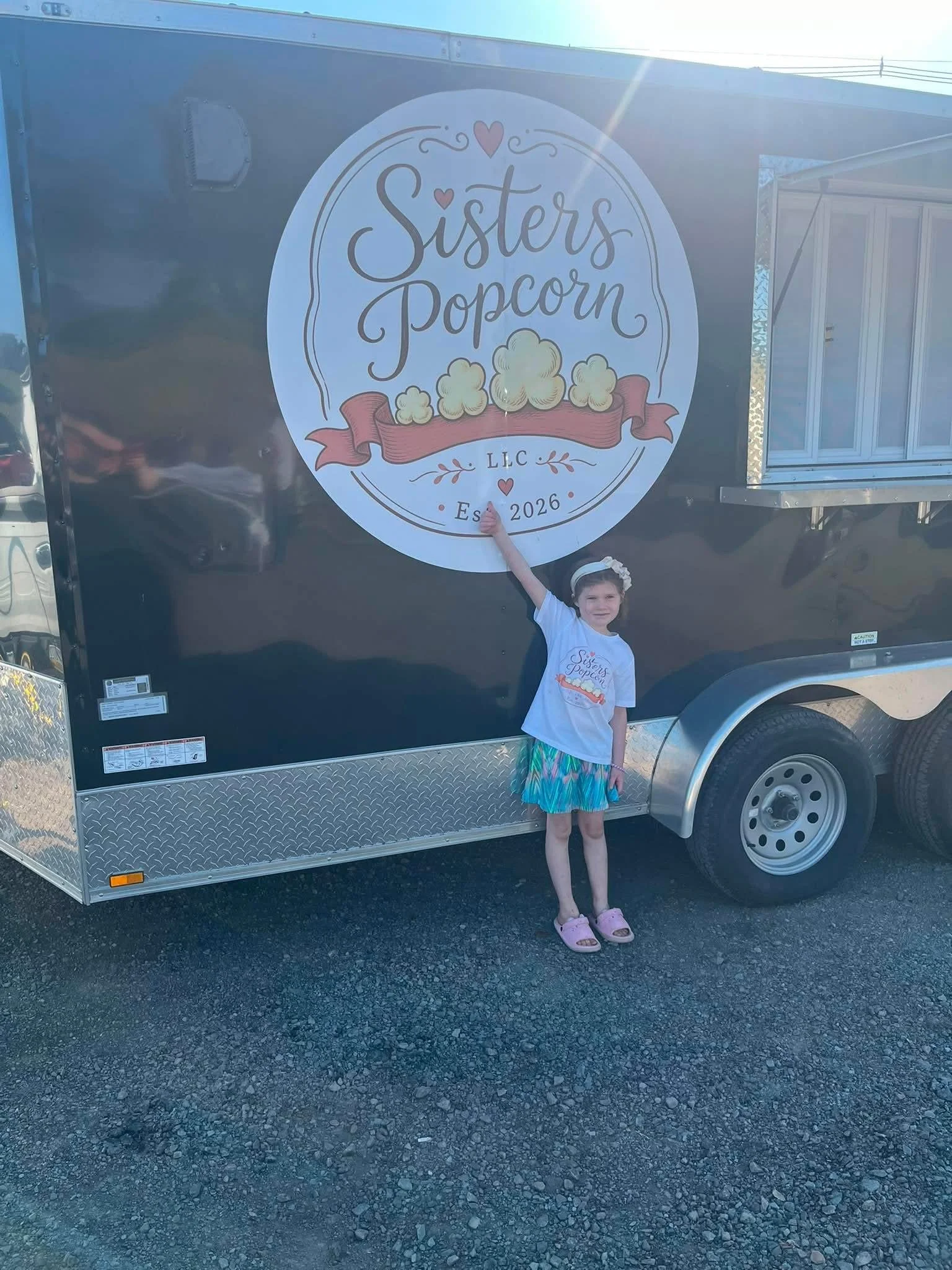 A young girl standing next to a black trailer with a large round sign that reads 'Sisters Popcorn LLC Est. 2026' with popcorn illustrations and decorative elements.