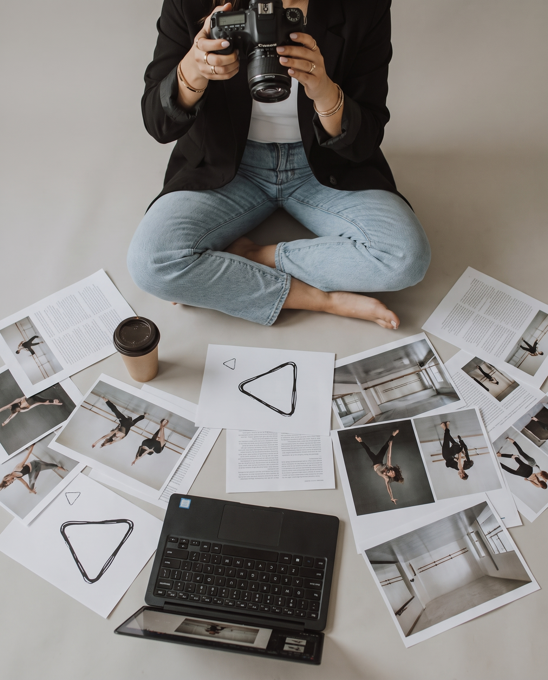A person sitting cross-legged on the floor, holding a Canon DSLR camera, surrounded by printed photographs of ballet dancers in various poses, a laptop, and a paper cup.