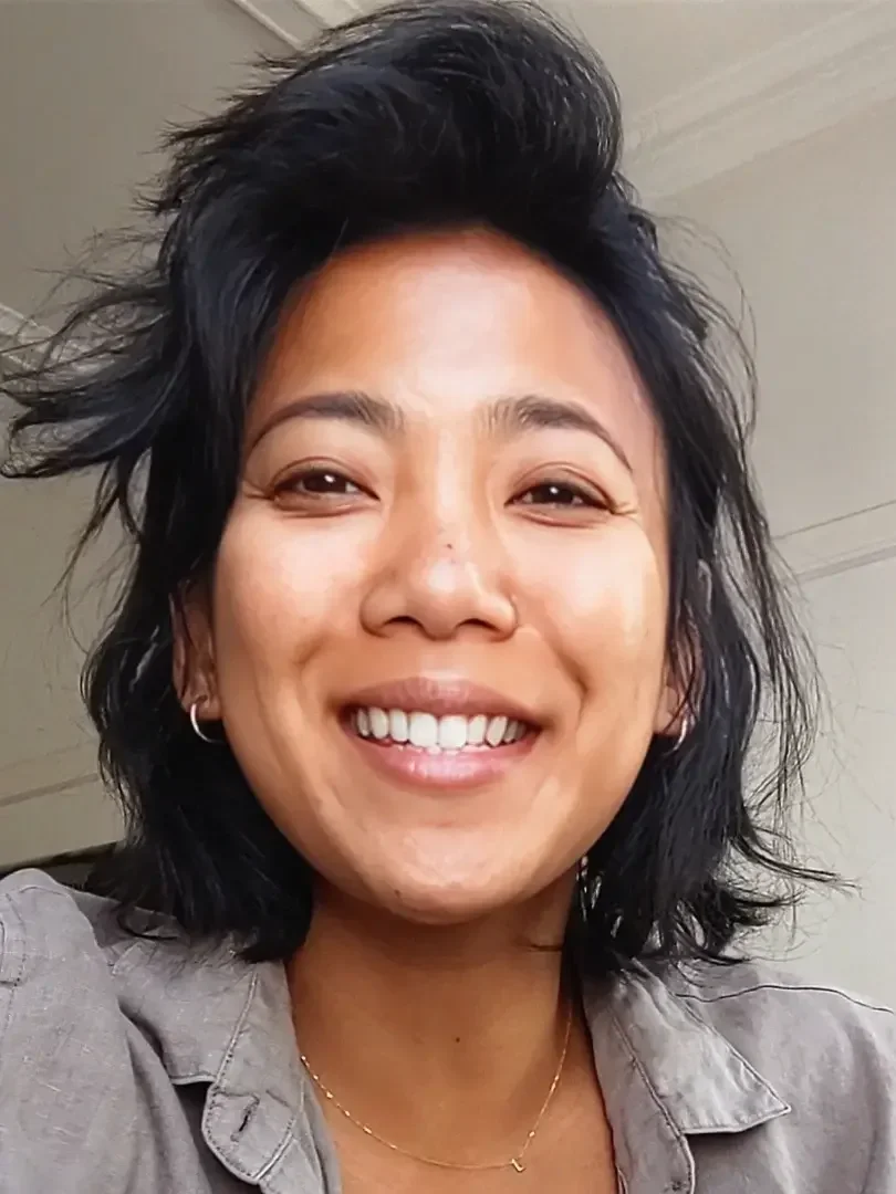 Close-up of a smiling woman with short, tousled black hair, wearing a gray shirt and small hoop earrings.