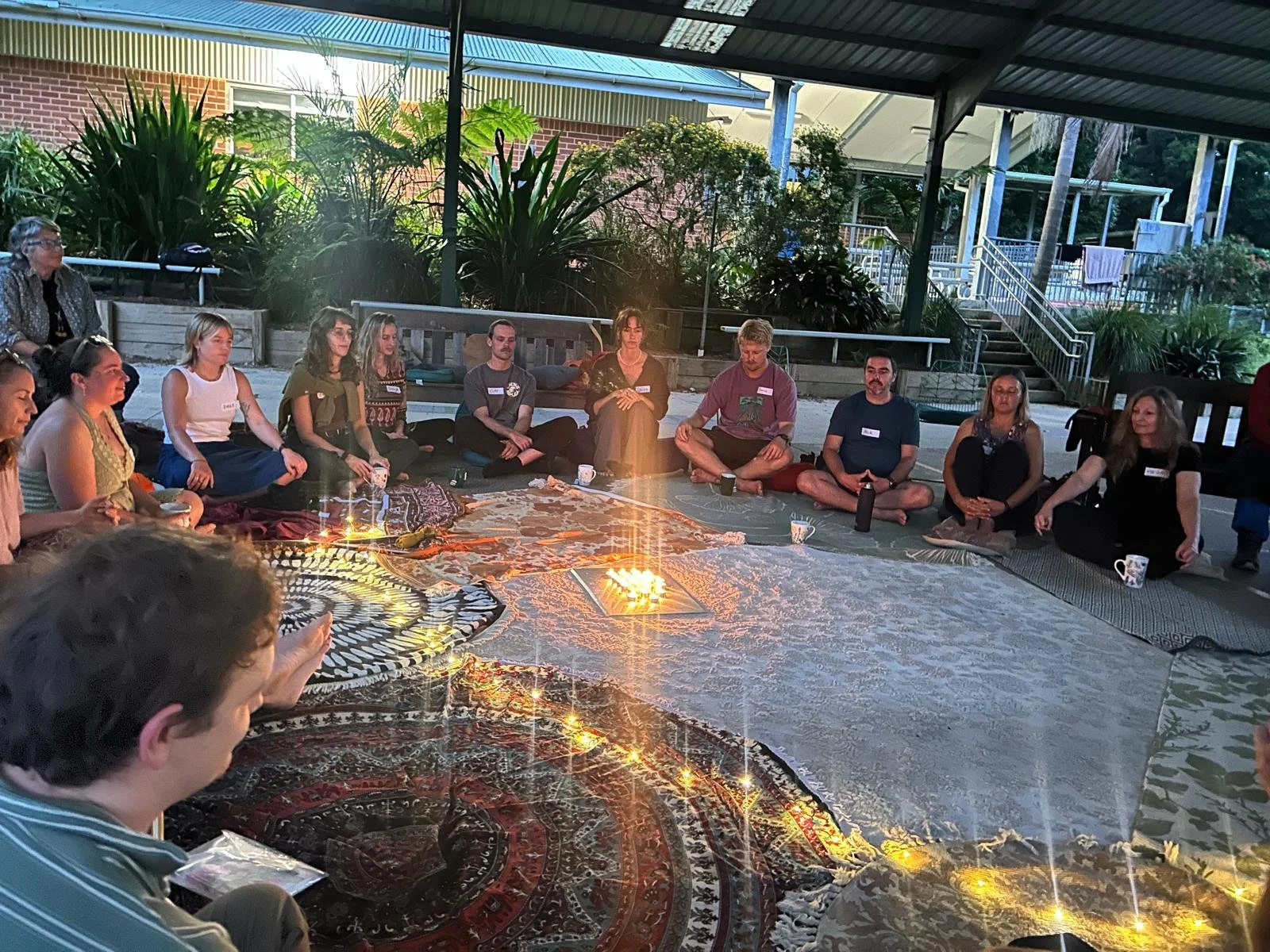 People sitting in a circle on rugs and blankets, participating in a group meditation or mindful gathering outdoors, illuminated by string lights with trees and a building in the background.