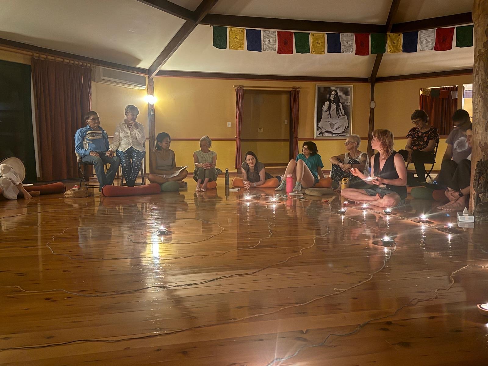 Group of women sitting on cushions and chairs in a circle on wooden floor, candles and water bottles, with prayer flags overhead, in a room with warm lighting and a framed black-and-white photo of a woman on the wall.