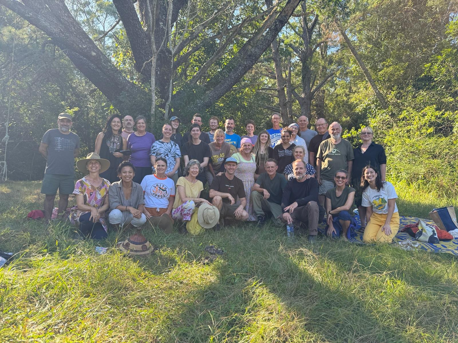 Group of people outdoors in a park, sitting and standing in front of large trees, smiling for a photo.