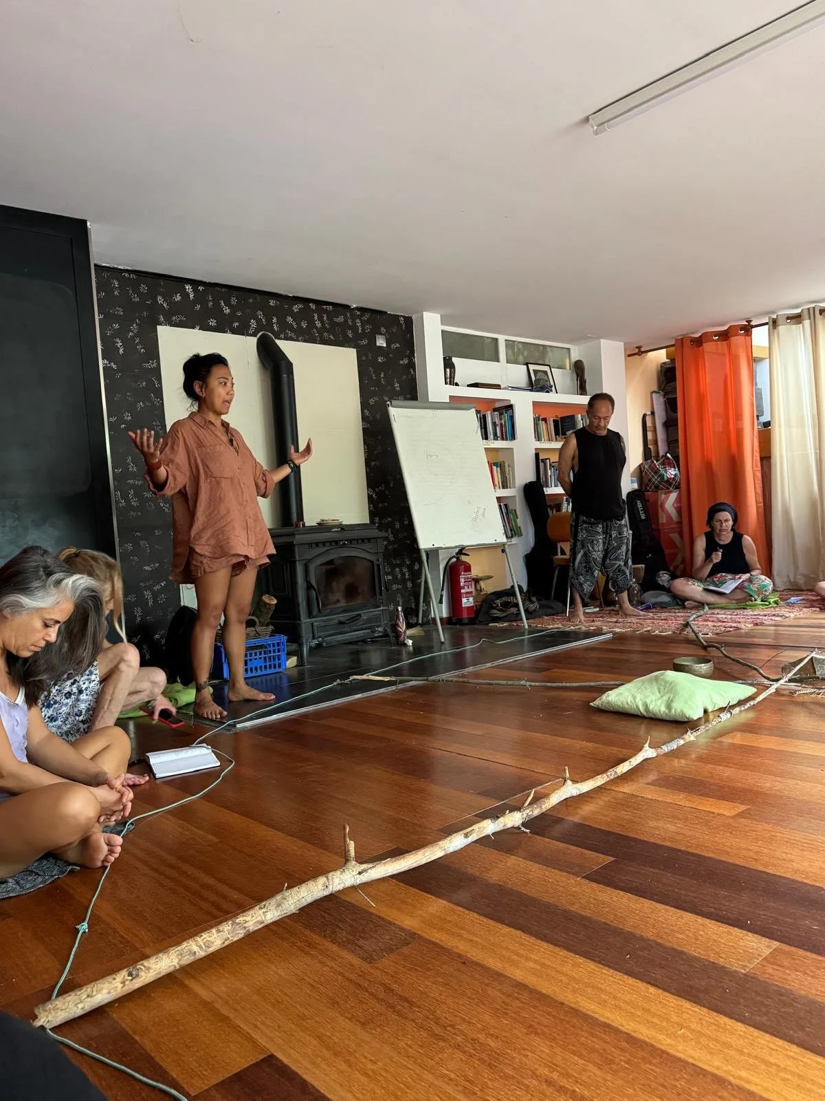 People sitting on the floor listening to a speaker in a living room. The woman standing is speaking, gesturing with her hands, wearing a brown shirt and shorts. Others are seated cross-legged on the floor, some taking notes or listening. The room has wooden floors, a whiteboard, bookshelves, and a firewood stove.