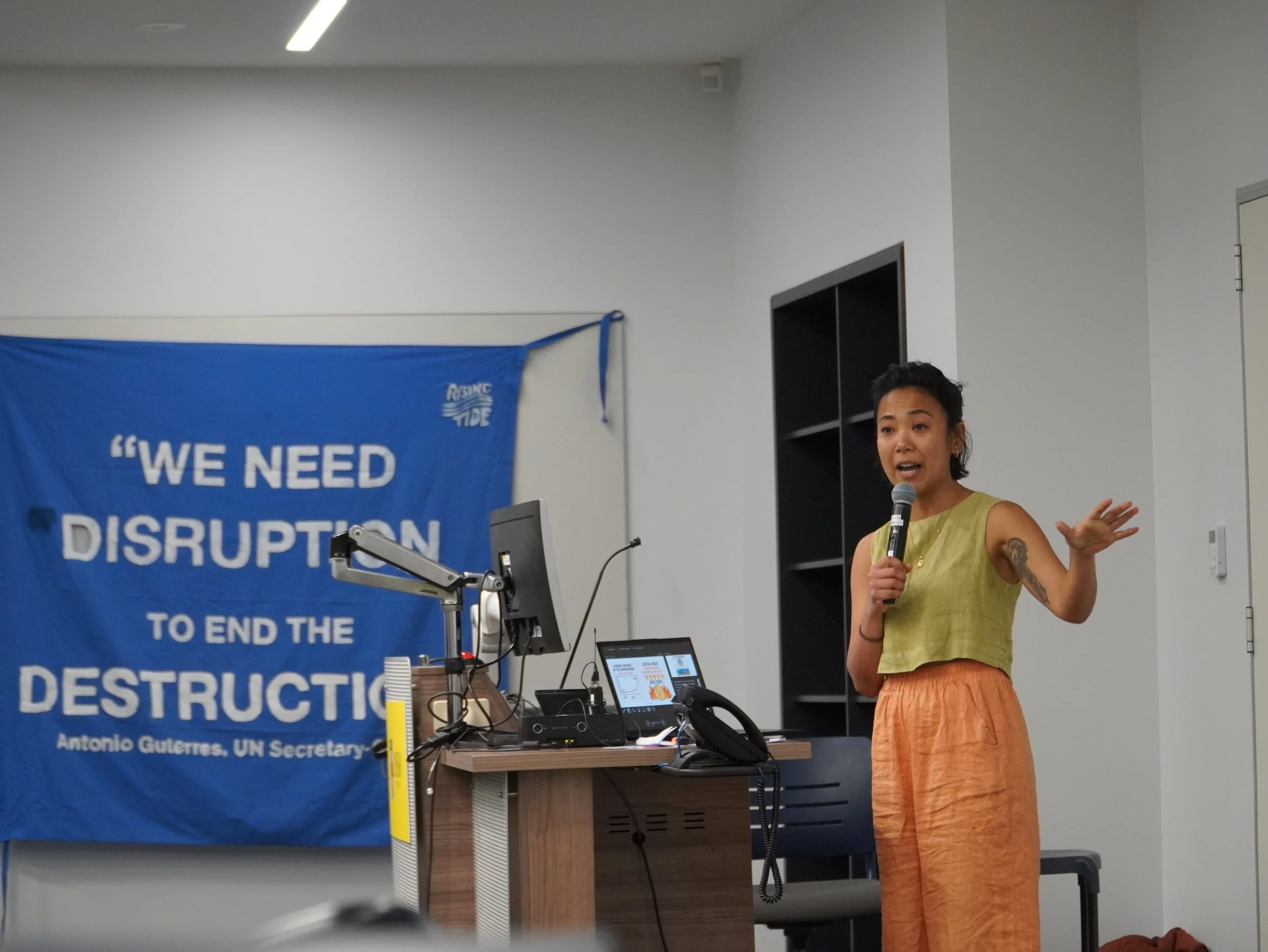 A woman speaking into a microphone during a presentation, standing next to a blue banner that reads 'We Need to End the Destruction.' She is wearing a yellow sleeveless top and orange skirt, with a tattoo visible on her left arm. The room has white walls, and there is a computer monitor and other electronic equipment on a desk in front of her.