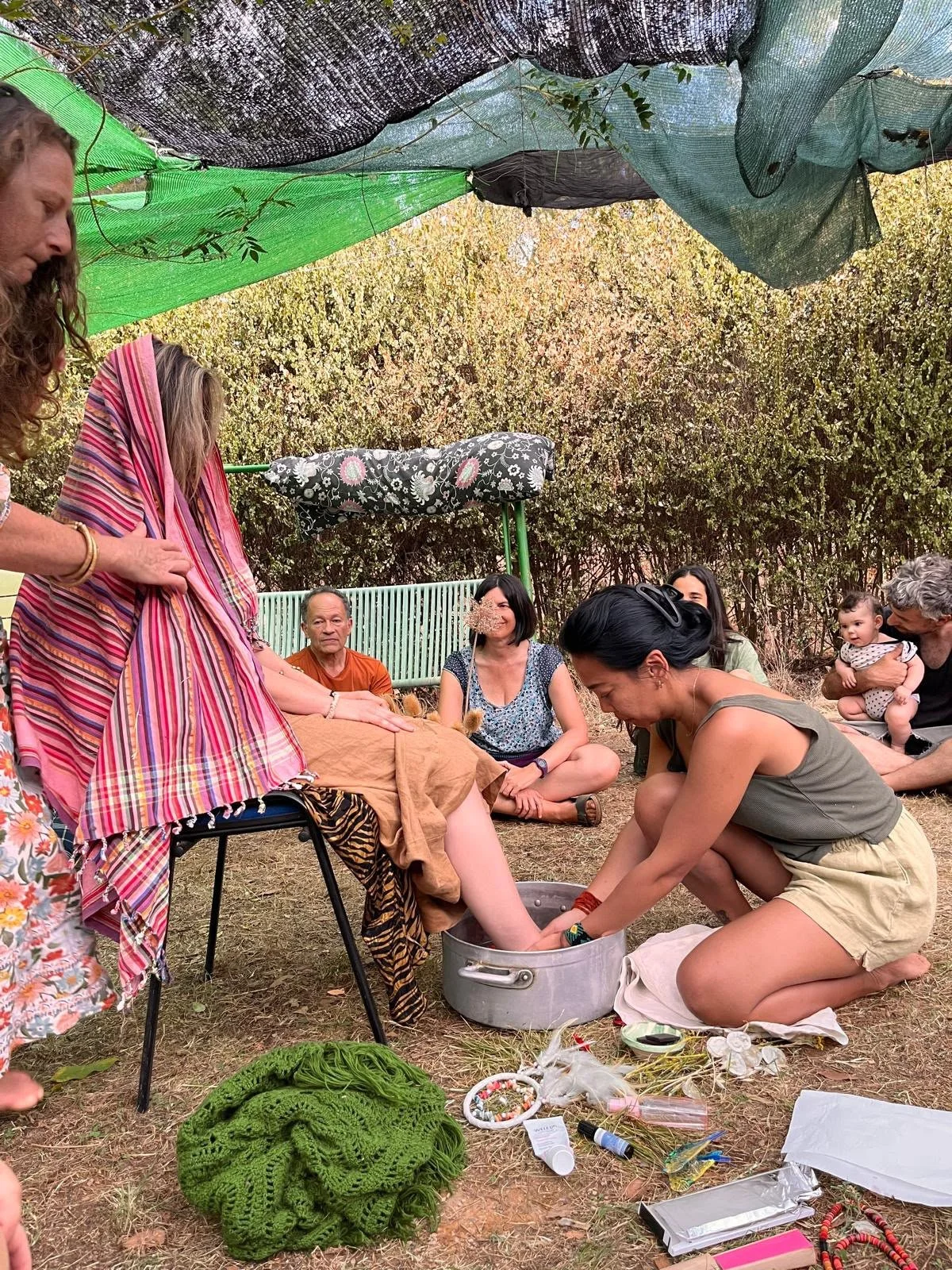 People participating in a spiritual or healing ceremony outdoors, with some sitting on the ground and one person massaging a woman's feet in a metal basin, under a colorful shade cloth with trees in the background.