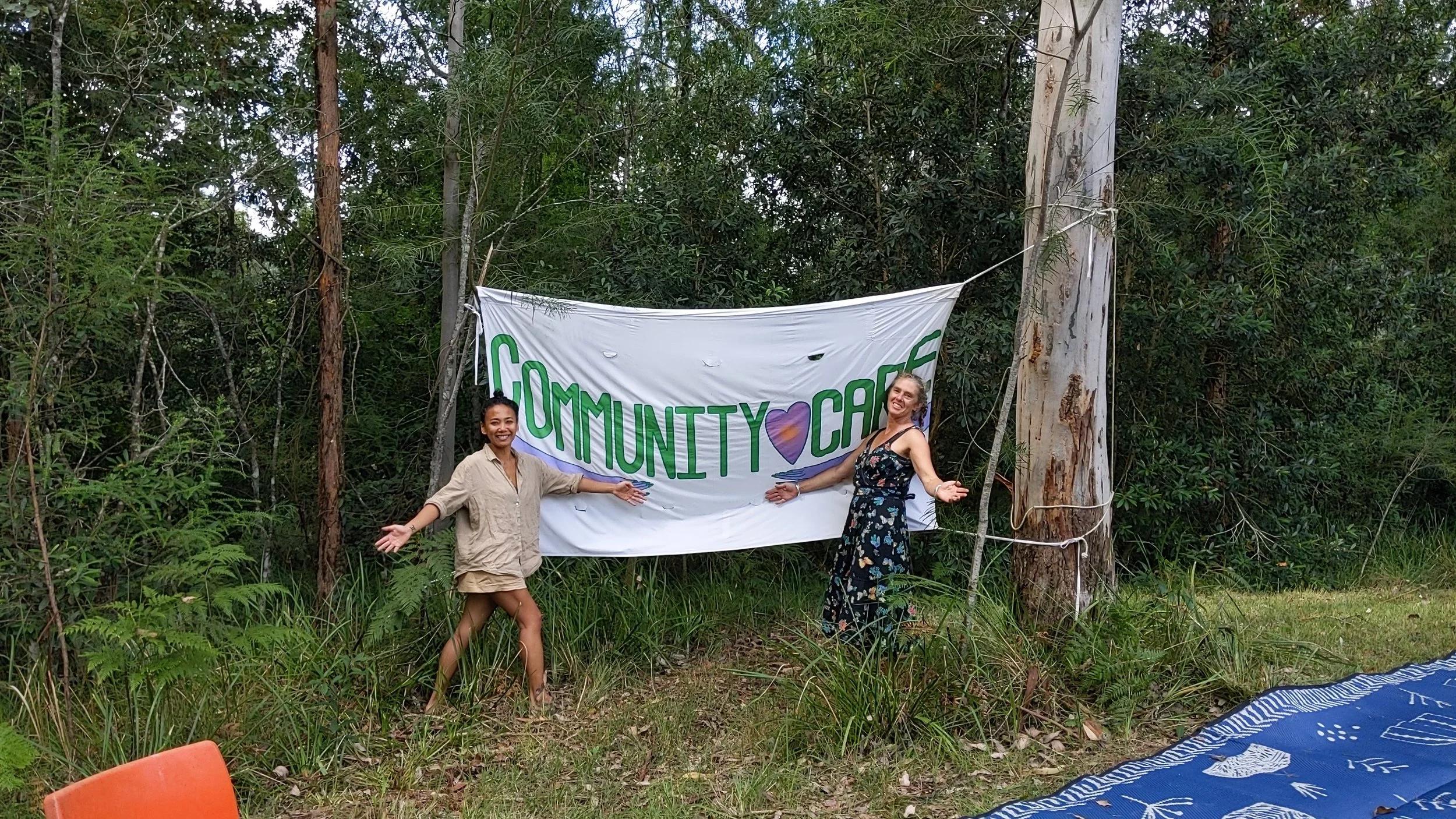 Two women smiling and posing in front of a white banner with green and purple writing that says 'Community Care,' in a forested outdoor setting.
