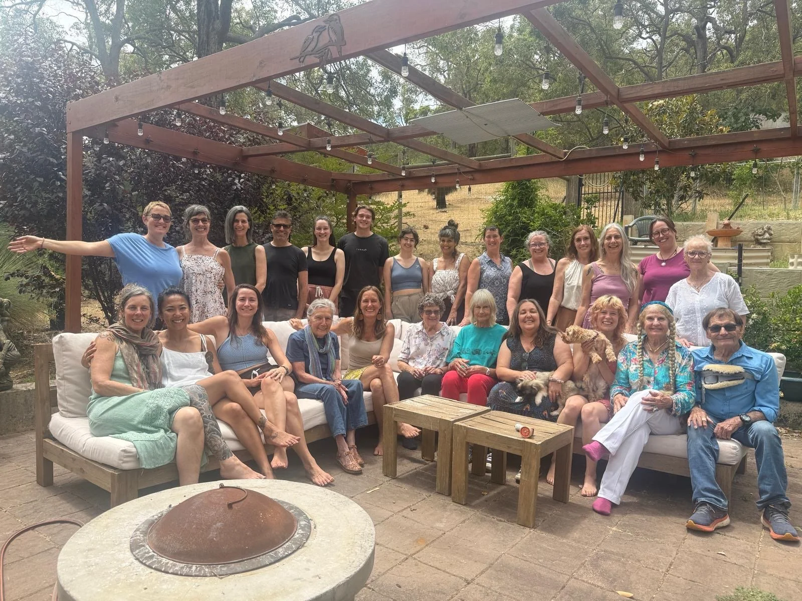 A large group of people gathered outdoors under a wooden pergola with string lights, posing for a group photo. Some are sitting on sofas, others standing behind. There are trees and greenery in the background, and a fire pit in the foreground.
