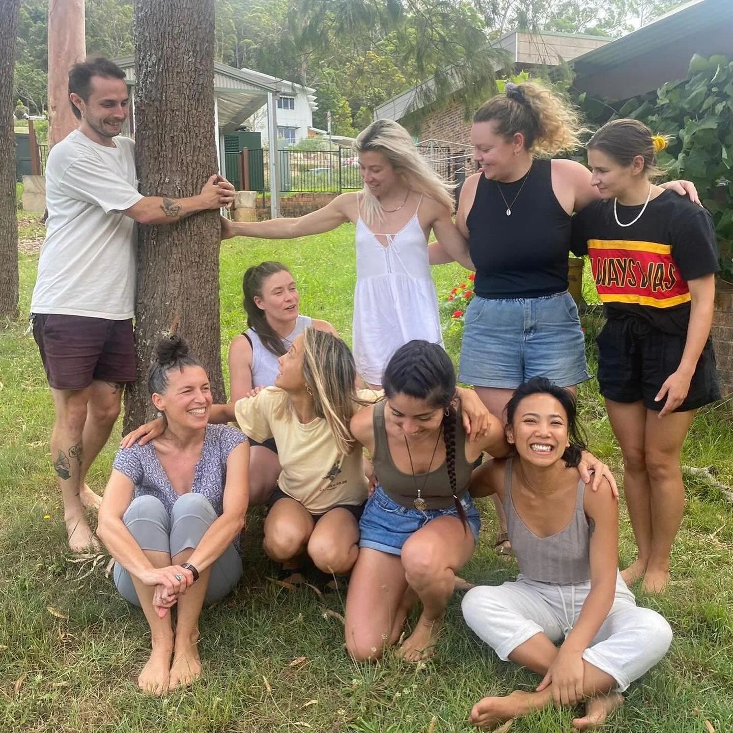 A group of ten diverse women and men are gathered outdoors near a tree, smiling and engaging in playful poses. Some are sitting on the grass, while others stand around, touching the tree or each other, enjoying a joyful moment together.