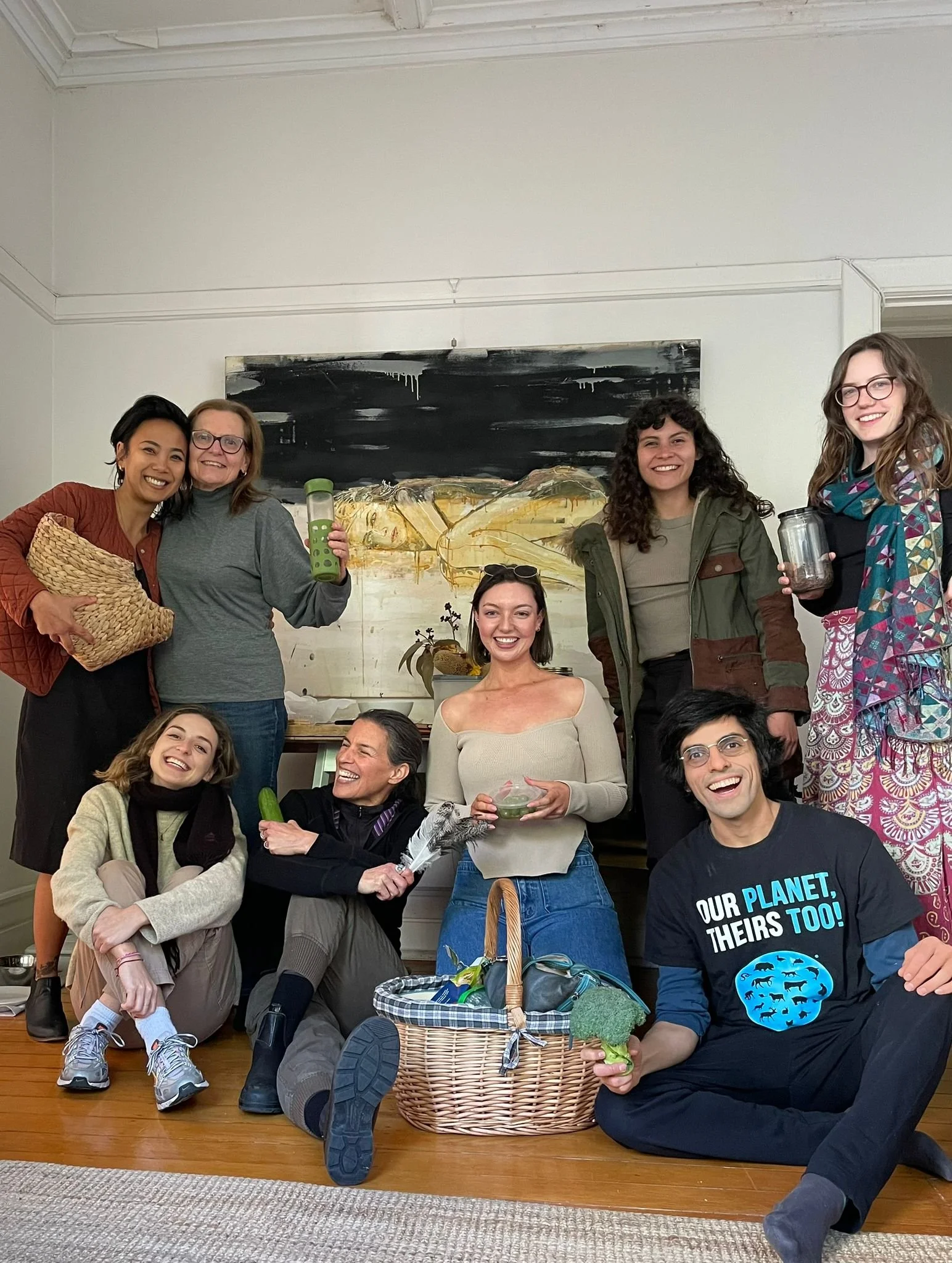 Group of ten people gathered indoors, some sitting and some standing, smiling and holding various objects including jars, a leaf, a feather, and a basket, with a large abstract painting in the background.