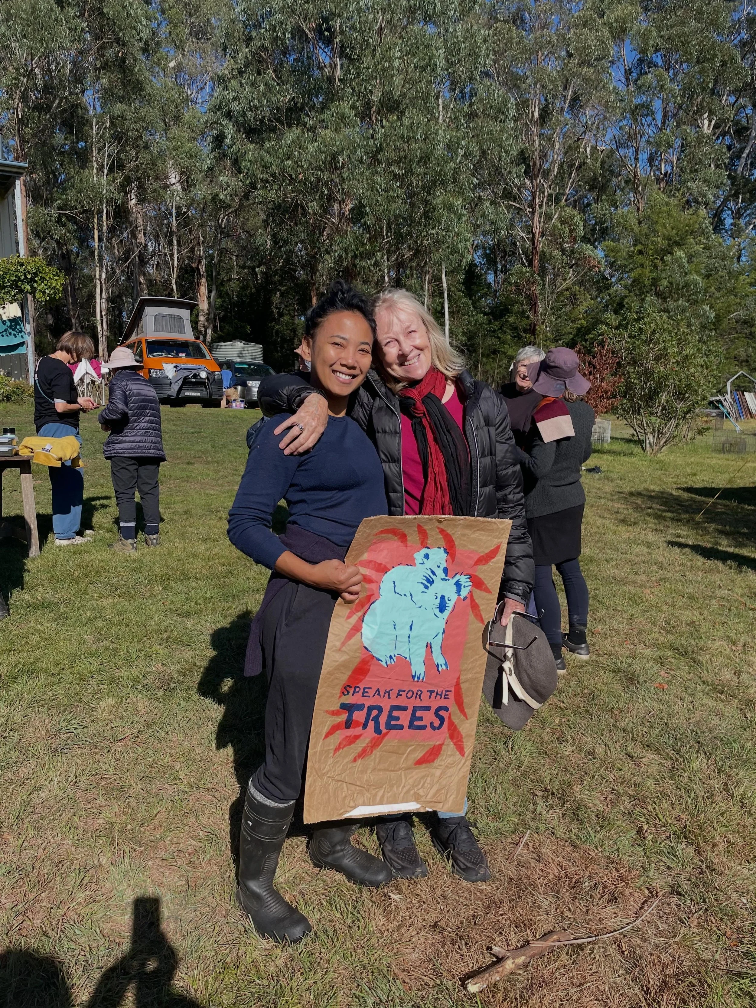 Two women smiling, one holding a 'Speak for the Trees' sign featuring a blue animal silhouette, outdoors on a sunny day with trees and other people in the background.
