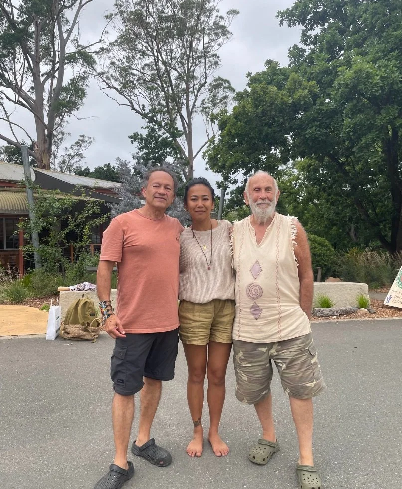 Three people standing outside on a cloudy day, with trees and a building in the background. They are smiling and posing for the photo.