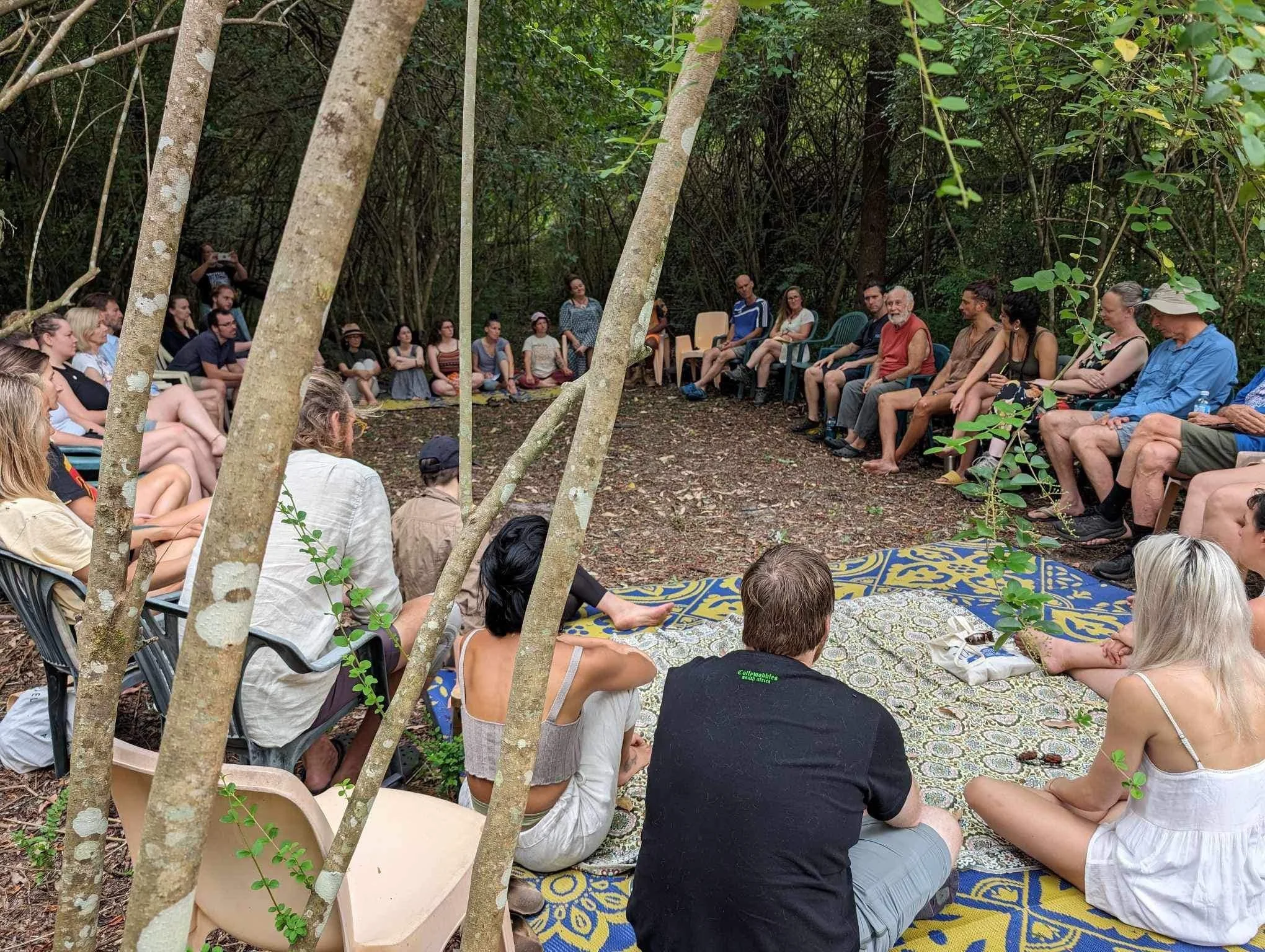 Group of people sitting in a circle in a wooded outdoor area, participating in a gathering or meditation session, with some seated on chairs and others on the ground, and a table with a cloth in the center.