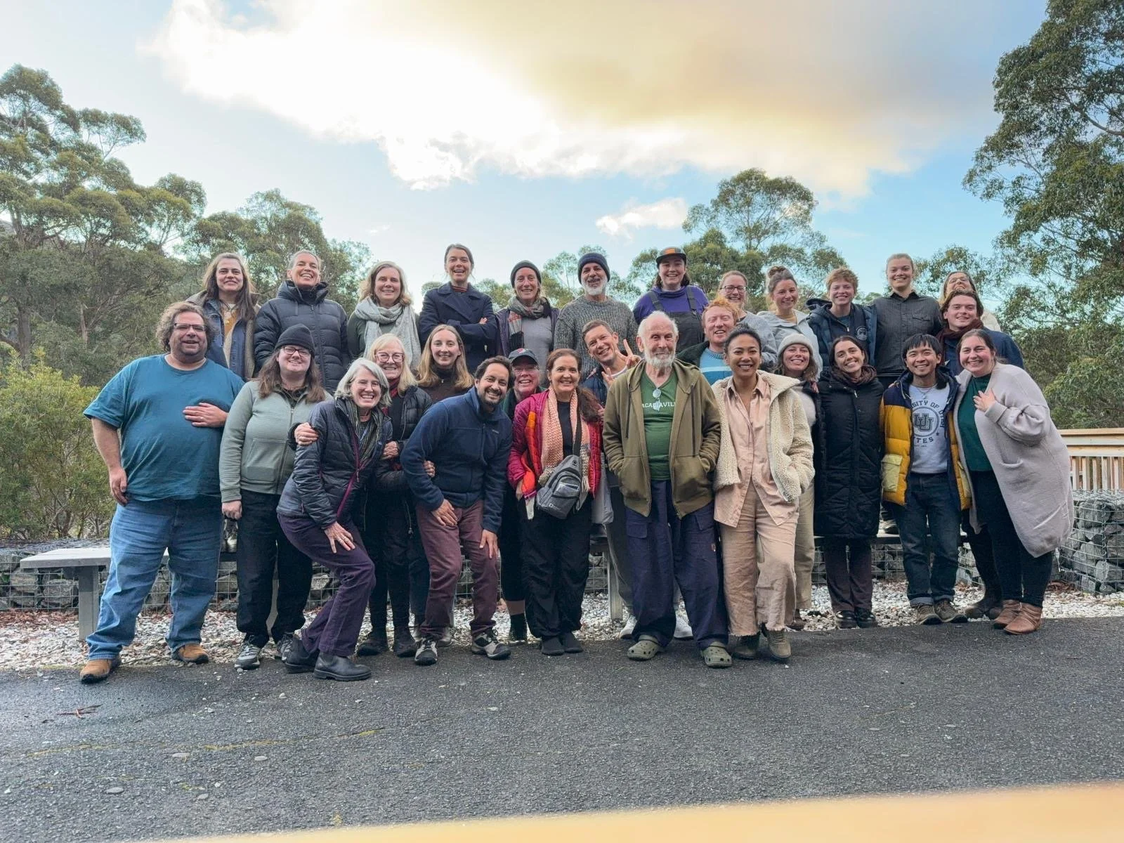 Group of smiling people outdoors on a cloudy day, standing in front of trees and a wooden railing.