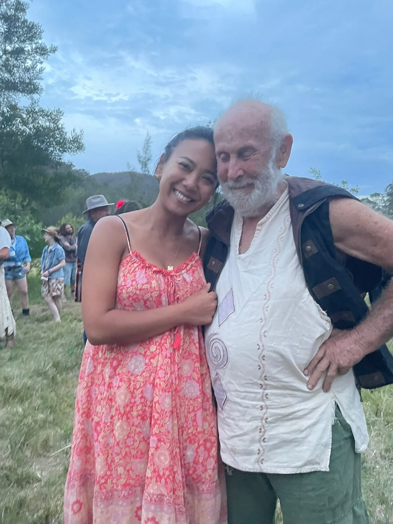Two people, a woman in a pink dress and an older man with a white beard, smiling and hugging outdoors during daytime, with a group of people in the background and a cloudy sky.