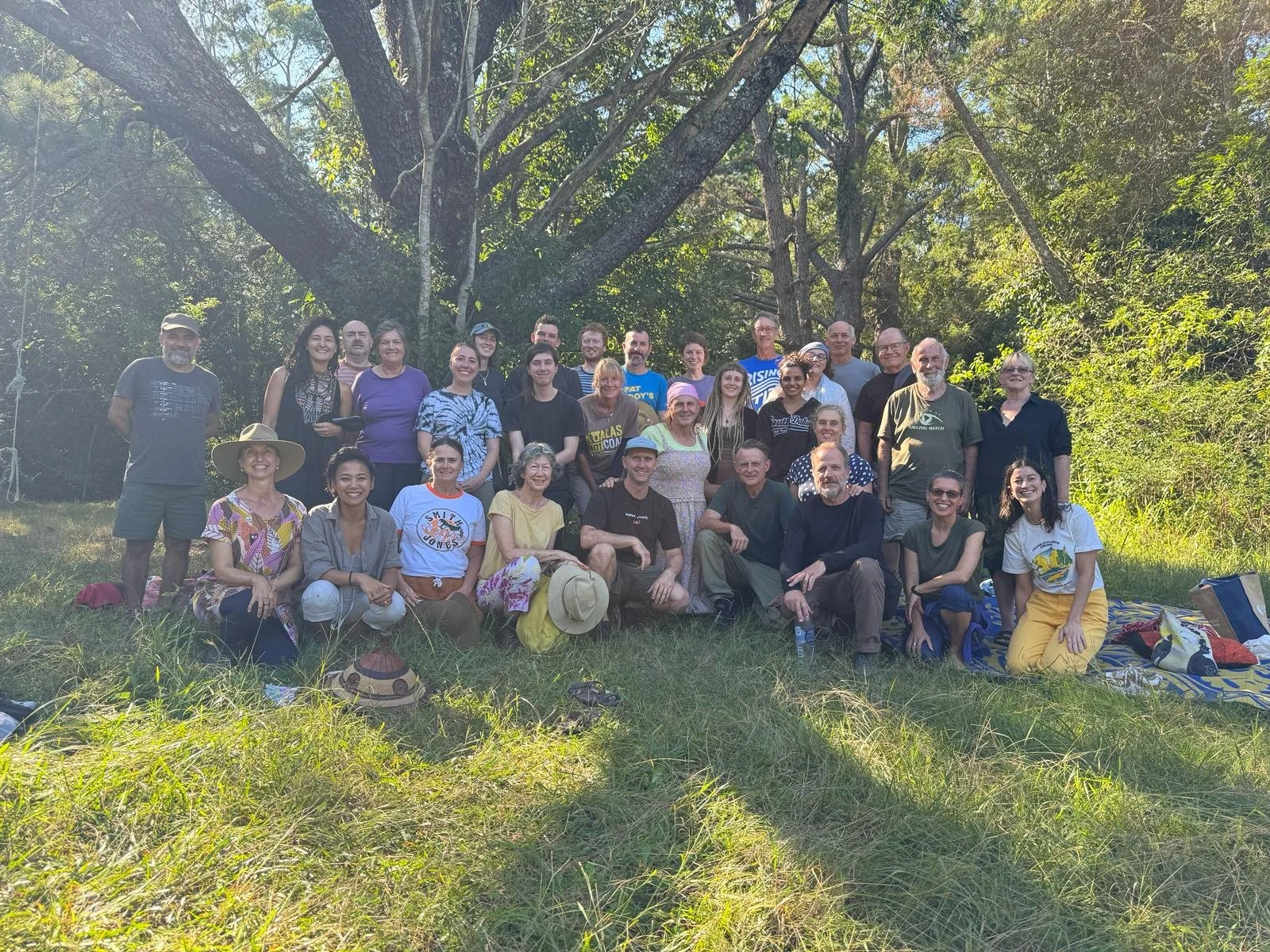 A large group of diverse people outdoors in a grassy area with trees in the background, posing for a group photo.