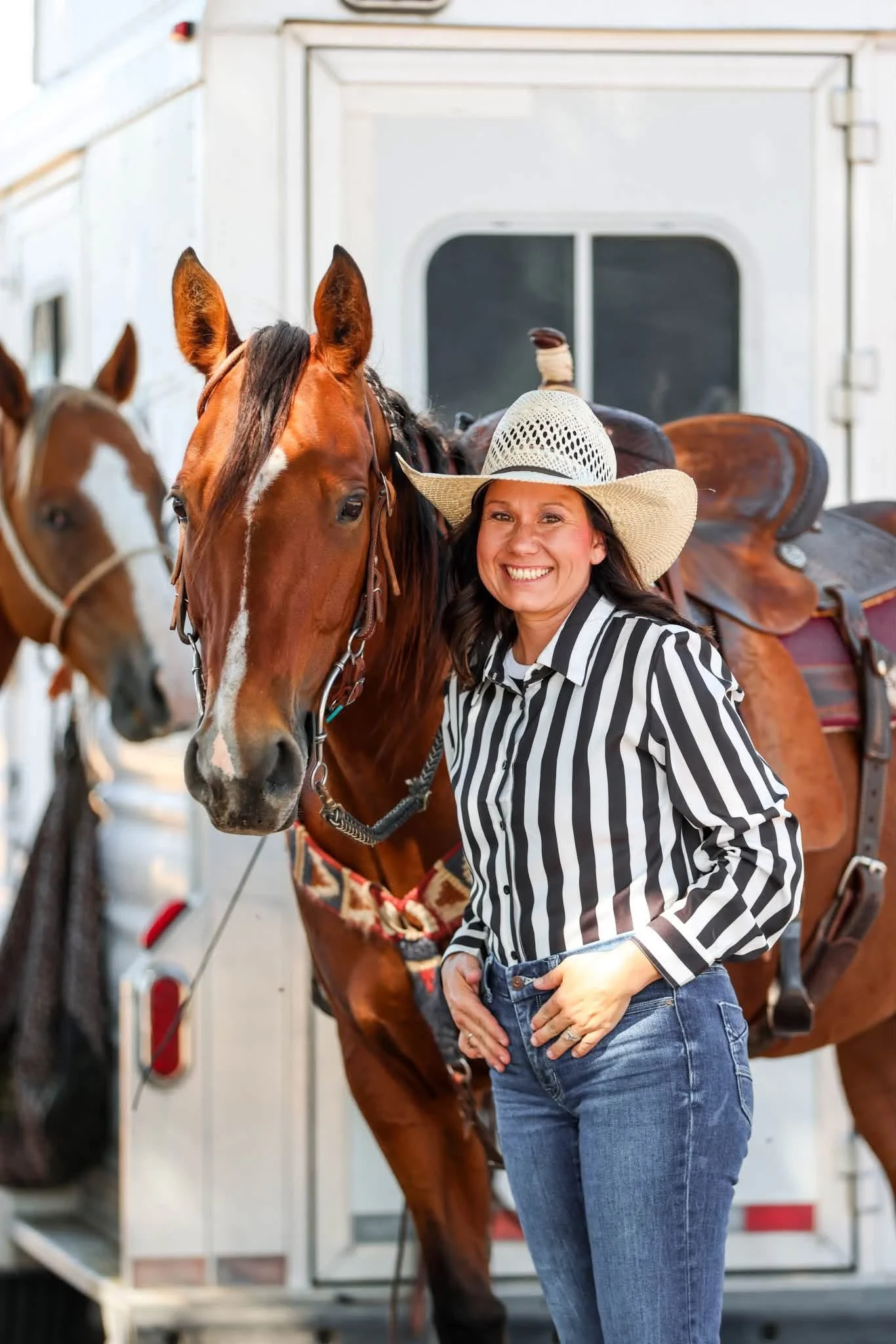 Woman in a black and white striped shirt and jeans smiling with a horse, standing inside a horse trailer with other horses in the background.
