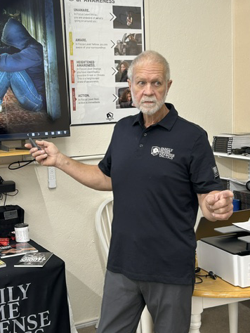 An older man with a beard wearing a navy polo shirt and gray pants, holding a remote control in a classroom or training room, standing next to a display screen. There are instructional posters on the wall and a table with various items.