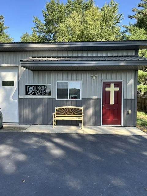 A gray building with a red door featuring a gold cross, a bench outside, a window, and a sign that reads 'Family Home Defense Tactics.' Trees are visible in the background.