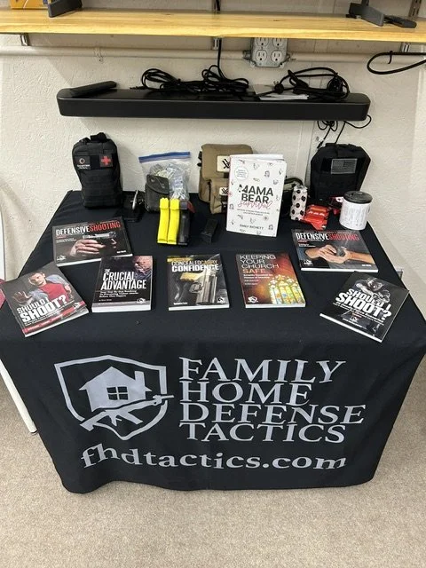 Table with family home defense tactics materials, including books, a first aid kit, flashlights, and self-defense items, covered with a tablecloth branded 'Family Home Defense Tactics'.