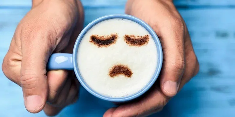 A person holding a blue coffee cup with a sad face made of cocoa powder on the foam.
