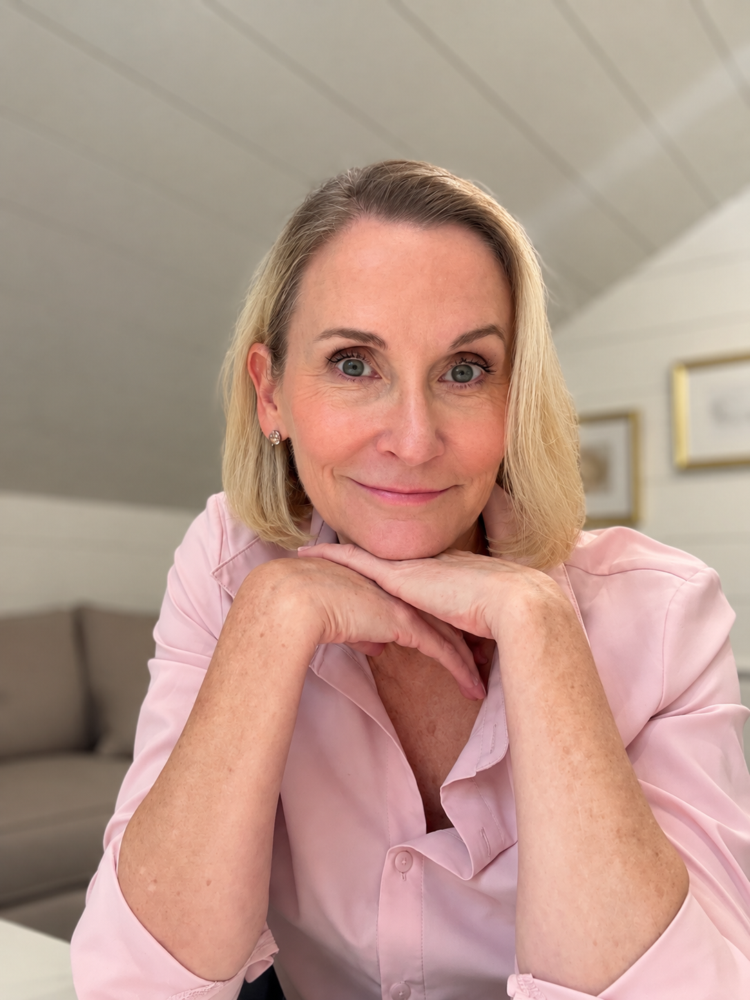 Close-up of a smiling middle-aged woman with blonde hair, wearing a pink blouse, resting her chin on her folded hands, in a cozy living room with beige furniture and framed pictures on the wall.