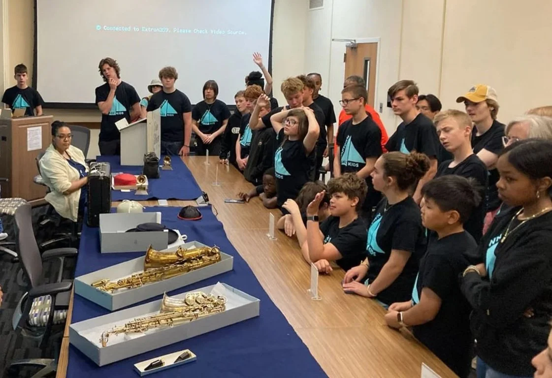 Group of children and teens standing around a table with musical instruments, including saxophones, in a classroom or conference room.