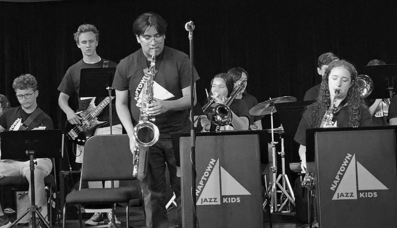 Young musicians playing jazz instruments during a performance, seen behind music stands with 'Naptown Jazz Kids' written on them.