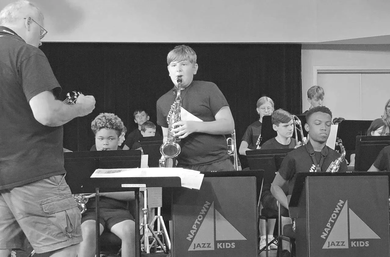 A young boy playing the saxophone during a musical performance on stage with a band of children, some holding instruments like a trombone and a guitar, in a venue labeled 'NAPTOWN JAZZ KIDS.' An adult man is standing to the side, possibly conducting or guiding the performers.