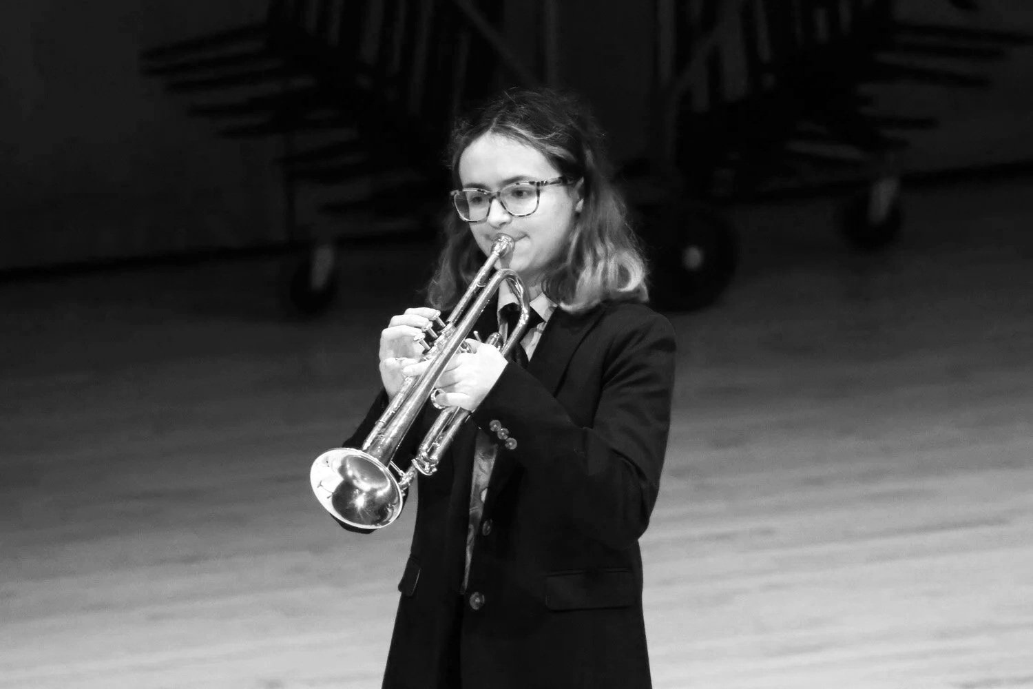 A young woman with glasses and wavy hair playing a trumpet on stage in black and white.