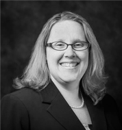Black and white professional headshot of a woman with shoulder-length hair, glasses, and a blazer, smiling at the camera.