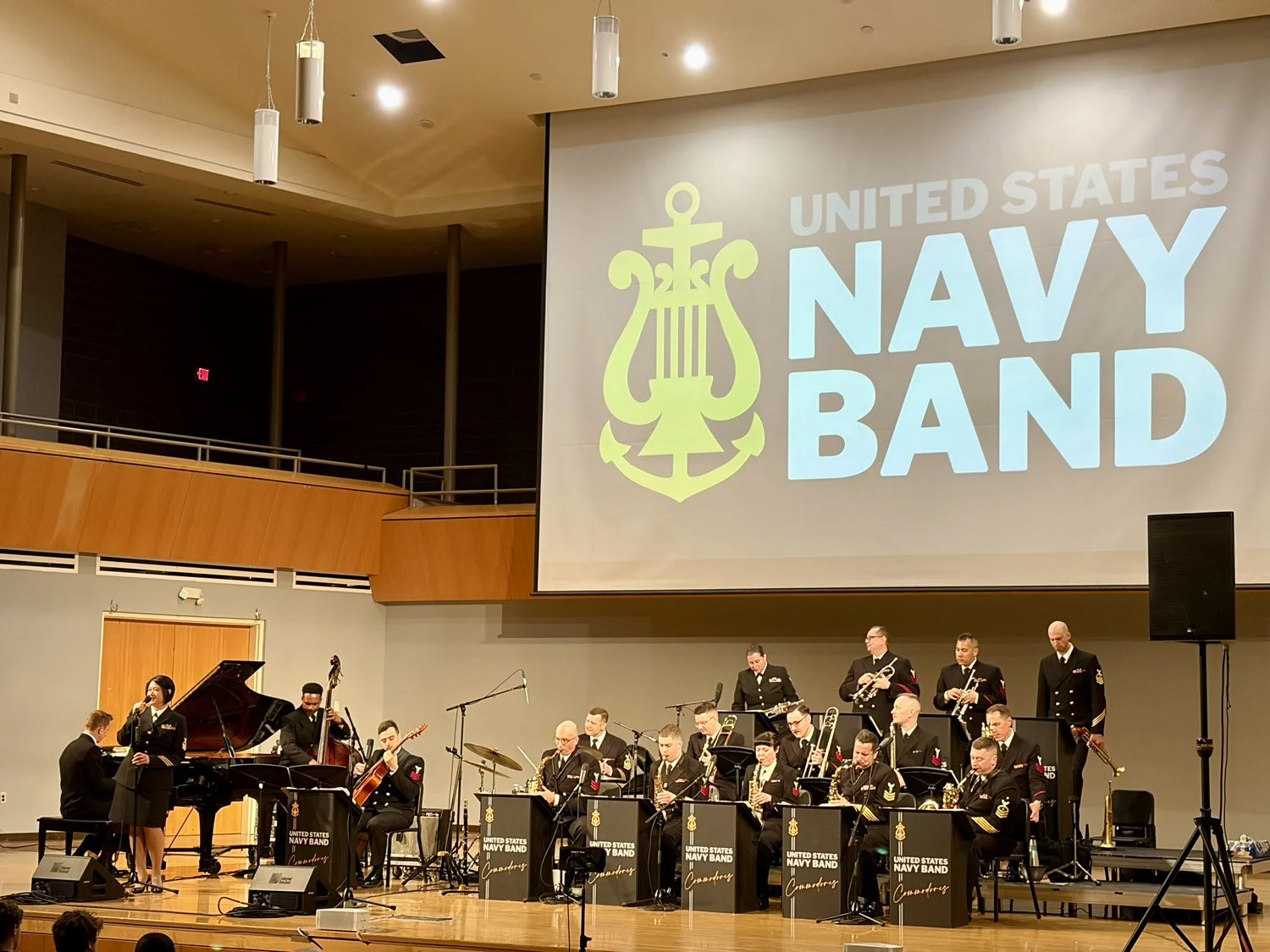 The United States Navy Band performs on stage in a concert hall, with a large screen displaying the Navy Band logo and name behind them.