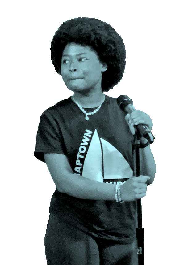 A woman with curly hair holding a microphone, wearing a T-shirt with a logo and text, standing against a black background.