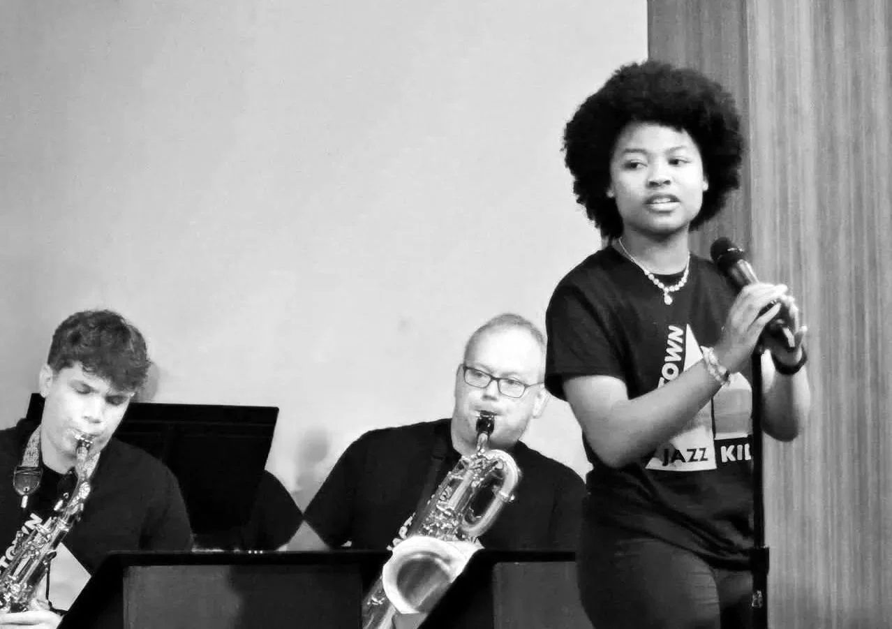 A black and white photo of a woman singing into a microphone with two men playing saxophones in the background at a jazz performance.