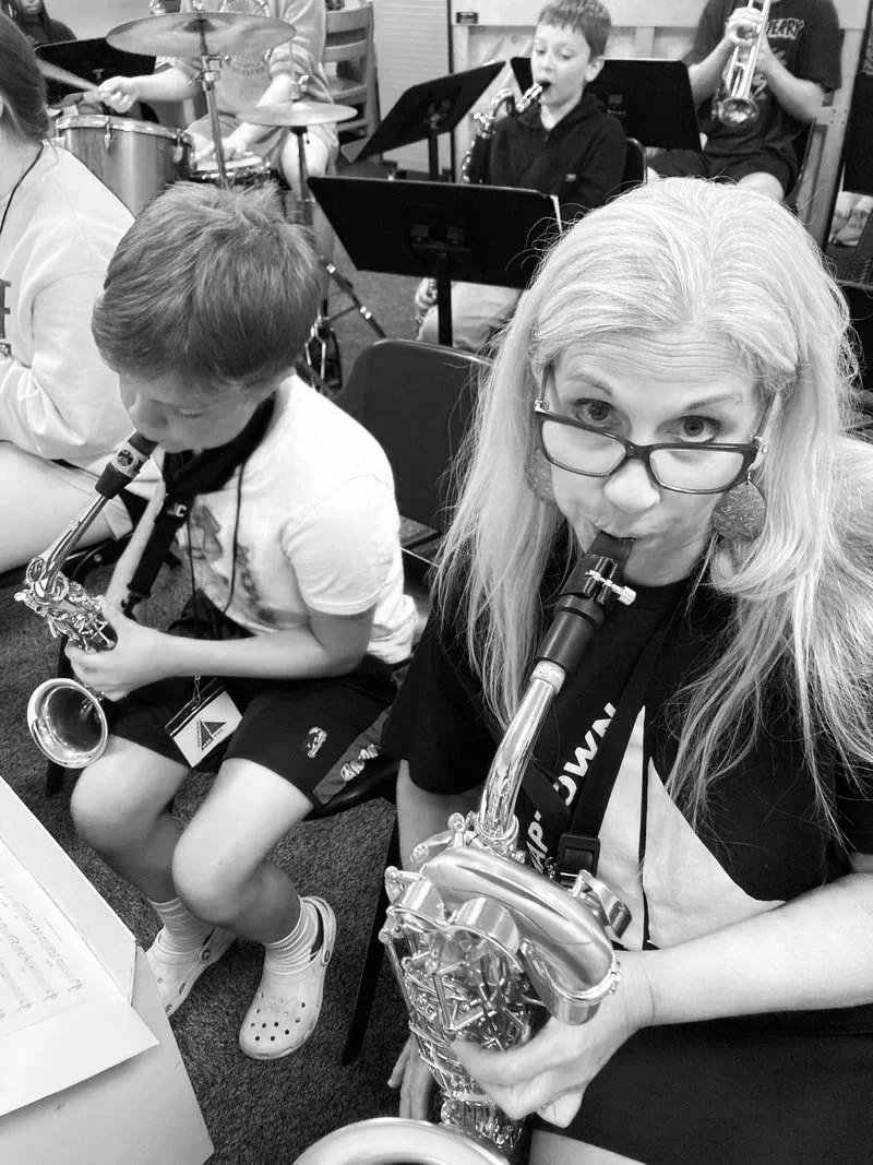 Adult woman and children in a band, playing saxophone and other instruments in a rehearsal room.