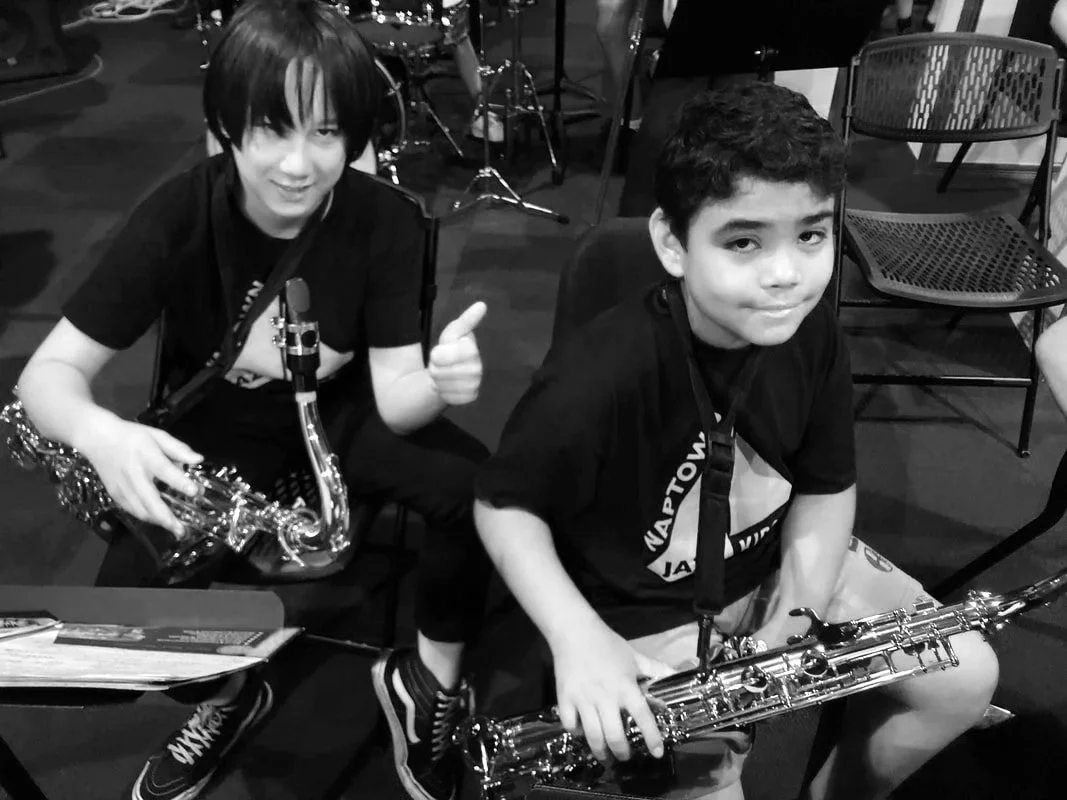 Two young boys sitting with saxophones, one giving a thumbs-up, in a music practice space with chairs and drums in the background.