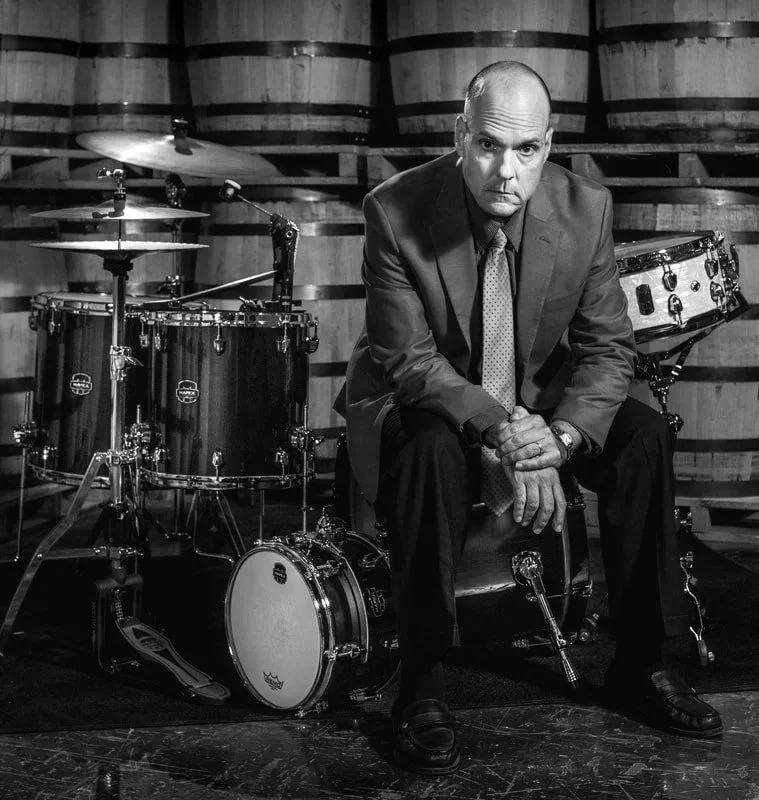 Man in a suit sitting on a stool in front of drums, in a cellar or bar setting with barrels in the background.