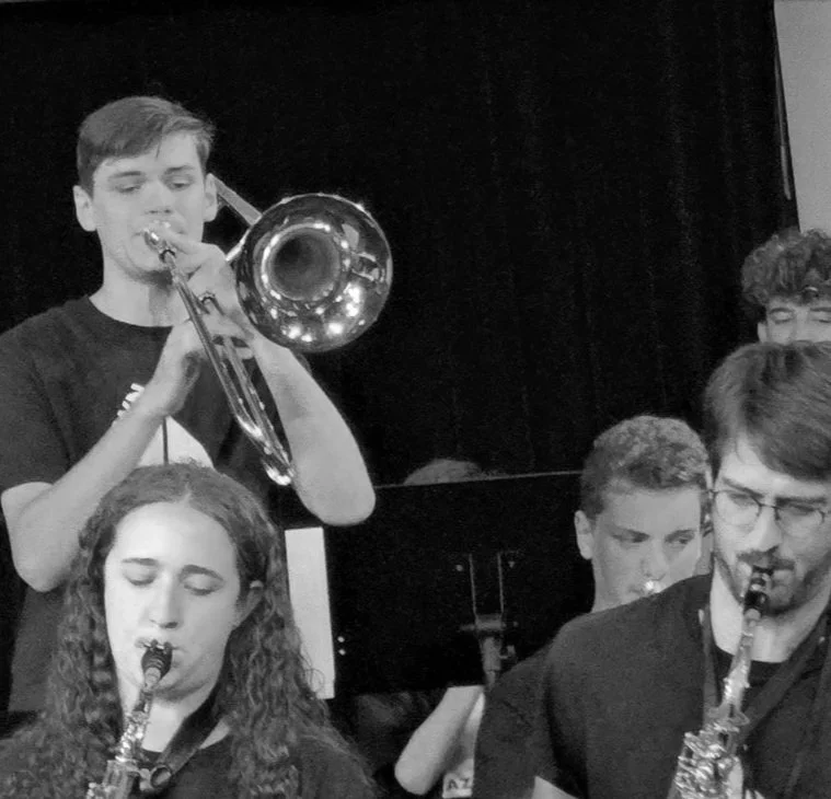 Black and white photo of young musicians playing wind instruments in a band rehearsal or performance, including a girl with curly hair playing a clarinet, a boy with a trumpet, and another boy with glasses playing a saxophone.