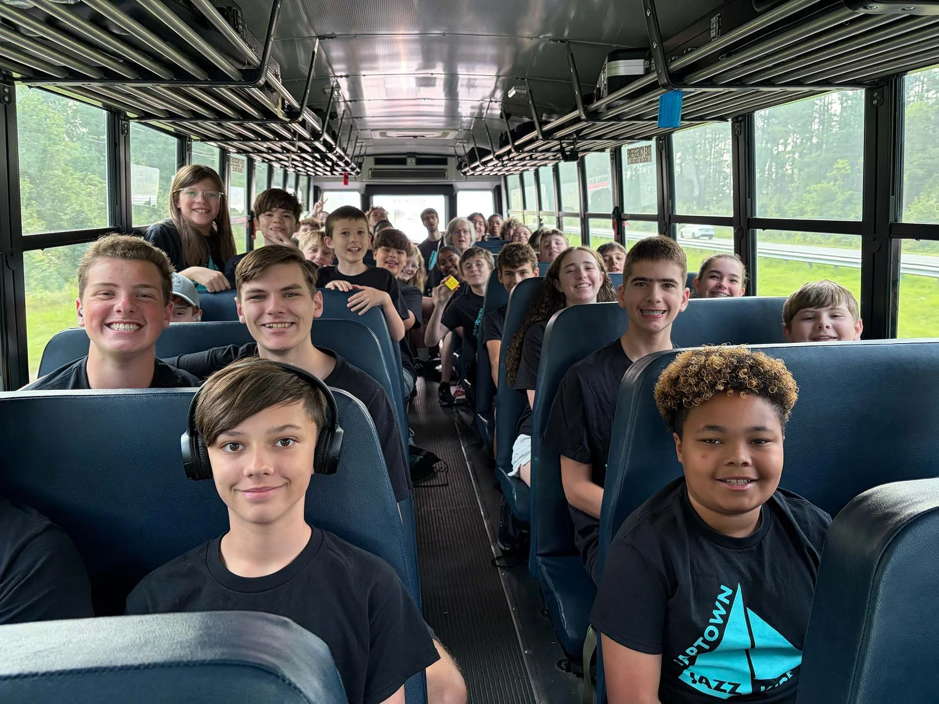 Young students, both boys and girls, sitting on a school bus, smiling. Some are wearing headphones, and they are looking forward or at the camera. The bus has large windows showing trees and greenery outside.