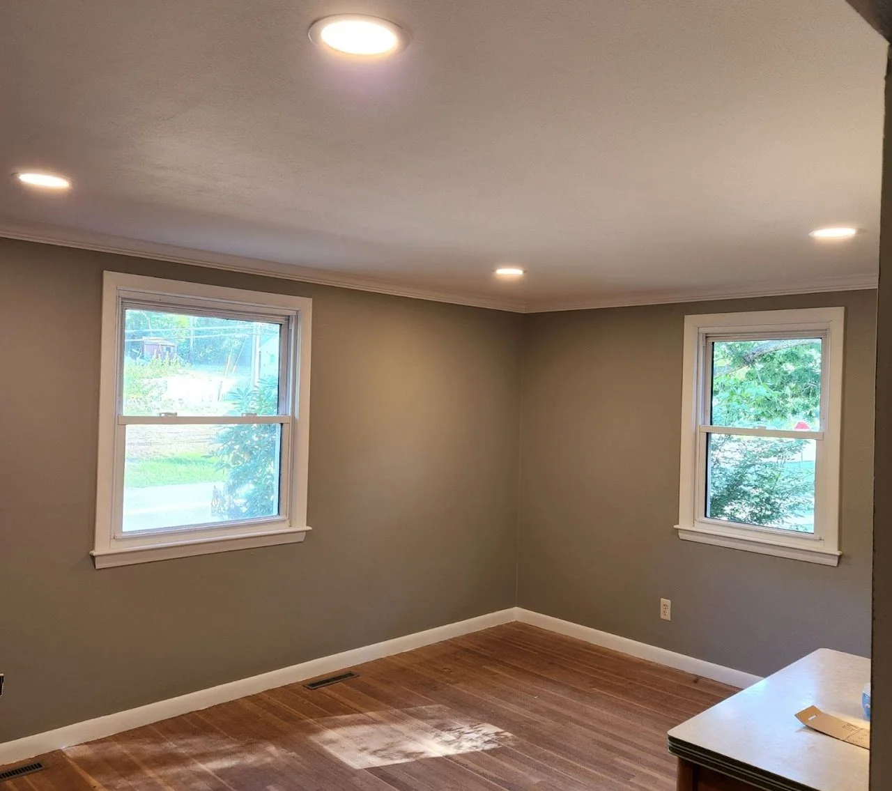 Empty room with two windows, green trees outside, hardwood floor, and recessed ceiling lights.