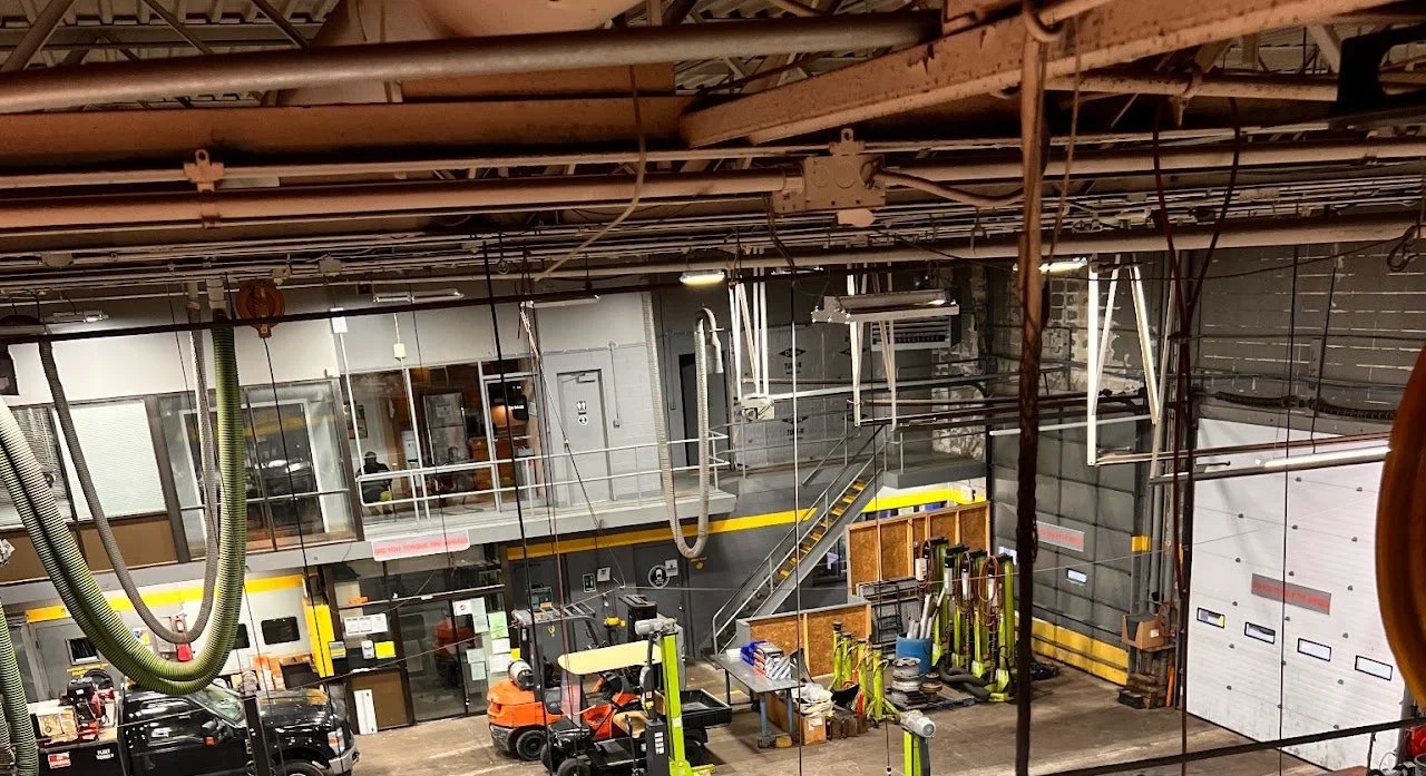 Indoor garage or workshop space with equipment, tools, and vehicles, including yellow and black safety features, stairs, and various machinery.