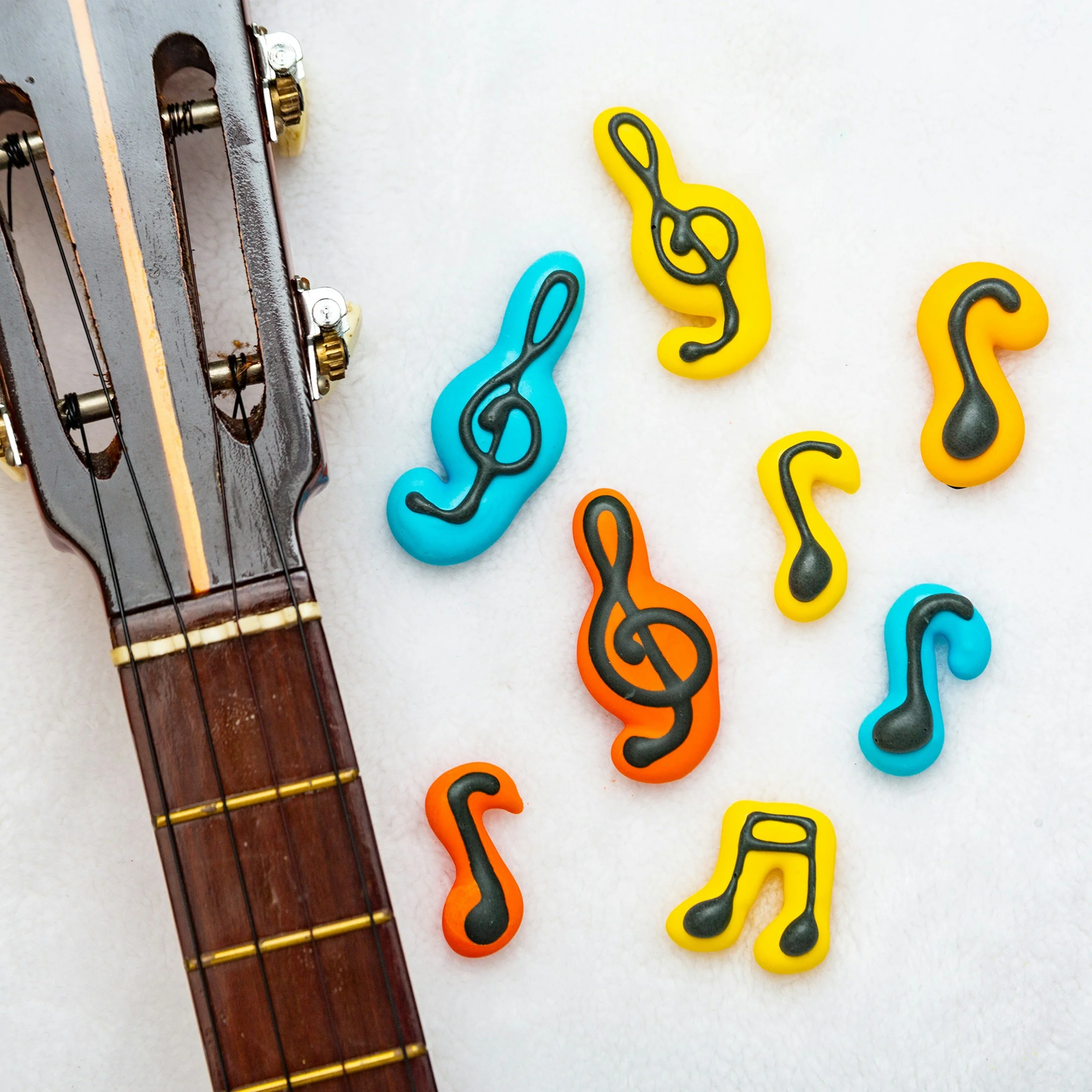 Close-up of a guitar neck with a metal tuning peg on the left side, and colorful plastic key-shaped hooks in yellow, orange, black, and blue on the right side, arranged on a white background.